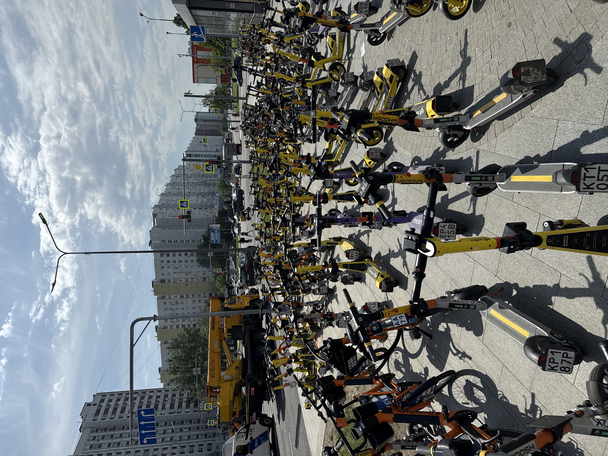 The image shows a large collection of rental electric scooters and bicycles parked on a paved area. The vehicles are predominantly yellow, with some orange bikes, and appear to be from various rental companies like Whoosh. In the background, there are apartment buildings, a street, and a yellow crane. The sky is partly cloudy. The location is in Moscow, Russia, given the license plate markings and the brand names written in Russian. The scooters are parked closely together, indicating a designated rental zone. The image appears to be taken during the daytime, likely in a residential or commercial area.