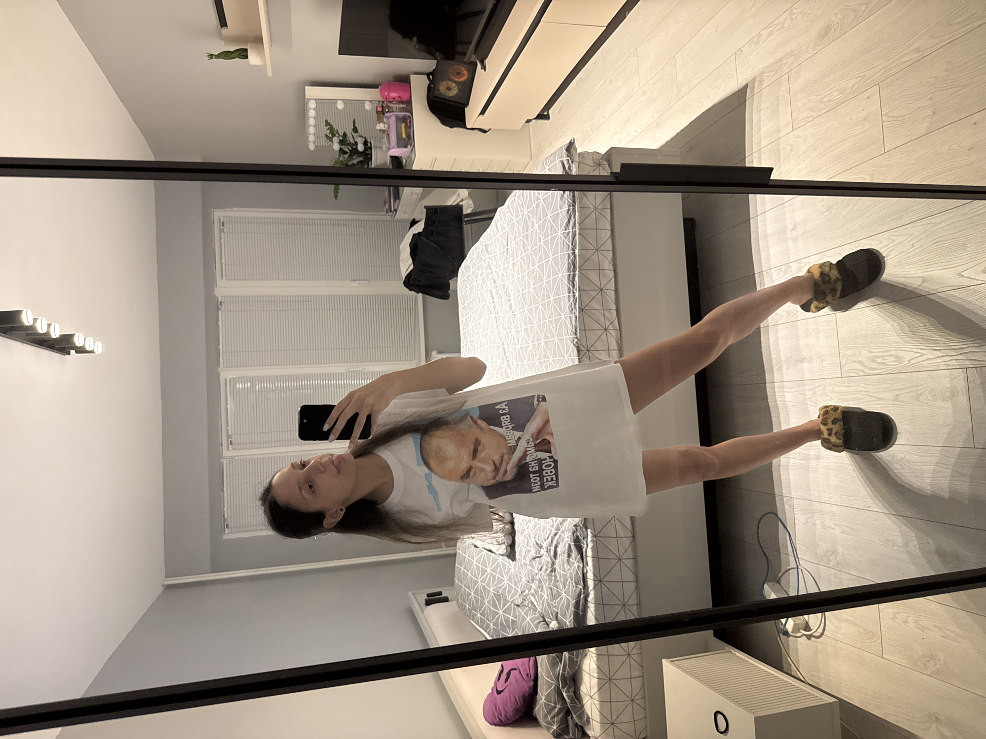 A young woman stands in front of a full-length mirror, taking a selfie with her phone. She's wearing a white t-shirt with a print of a man's face on it, and fuzzy slippers. The background reveals a bedroom with a bed covered in a geometric-patterned duvet, a TV unit, blinds on the windows, and other furniture. While the exact location within Sofia, Bulgaria cannot be definitively determined from the image, the style of the interior suggests a modern apartment.