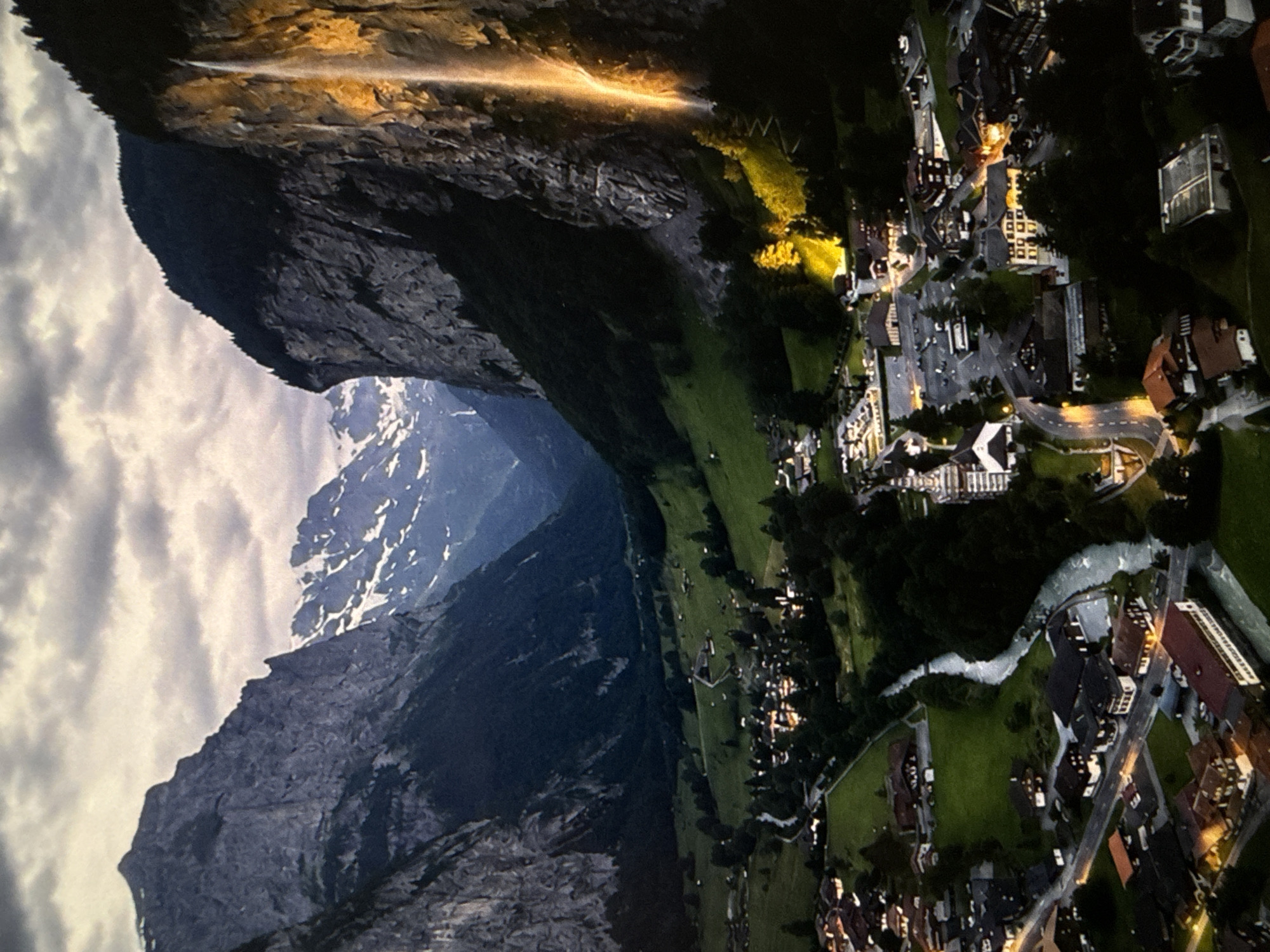 This is an aerial drone shot of the Lauterbrunnen Valley in Switzerland. The valley is framed by steep, dark grey cliffs on both sides. On the right side of the frame, a tall, thin waterfall cascades down the cliff face, illuminated by a golden light, possibly from artificial lighting or the setting sun. The waterfall appears to be the Staubbach Falls, one of the highest free-falling waterfalls in Europe.
Below the cliffs and waterfall, a lush green valley floor is visible. A small, illuminated village is nestled within the valley, with buildings clustered together and lights glowing from windows. A winding river flows through the village and the valley. In the background, more rugged mountains, partially covered in snow, rise into a cloudy sky. The overall impression is one of dramatic natural beauty, with the man-made village providing a sense of human habitation within the grand landscape. The lighting suggests it is either dusk or dawn, as the sky is overcast and the village lights are on.