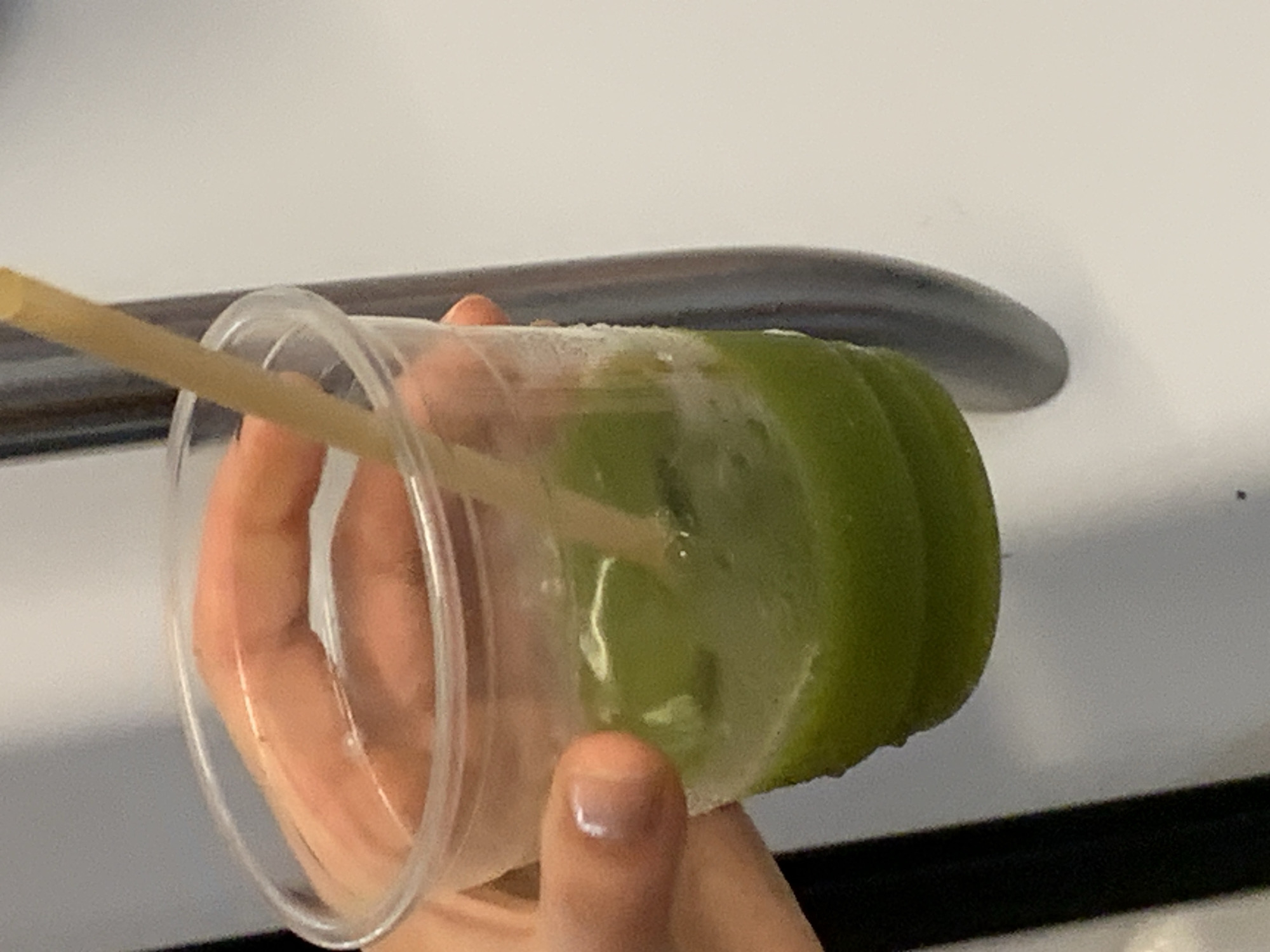 A hand holds a clear plastic cup filled with a green, icy beverage. A straw made of straw material sticks out of the drink. The cup is partially translucent, revealing the green contents within. The background is a white, slightly textured surface, possibly a wall or appliance, with a dark horizontal bar running across the lower portion. A curved metallic handle is visible at the top of the frame. The lighting suggests an indoor setting. There is no visible text in the image. The focus is on the drink and the hand holding it, suggesting the image was taken for personal or promotional purposes related to the beverage. The presence of ice in the drink and the overall cool tone of the beverage might imply a warm day or a desire for refreshment.
