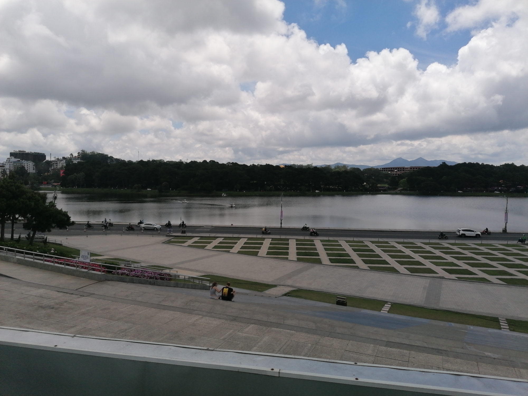 A medium-shot view depicts a man and woman sitting on a sloping concrete surface facing away from the camera towards a lake, likely situated in Da Lat, Vietnam. The couple is positioned near the lower center of the frame.
The foreground features a patterned concrete expanse with rectangular grass inlays. Beyond this, a road runs parallel to the lake. The lake itself has a smooth surface with distant boats.
The background showcases a forest line along the lake's edge, with buildings visible on the far left and mountains in the distance on the right. The sky is filled with a mix of white and gray clouds. Vehicles are observed moving along the road, and poles are situated along the lakeside.