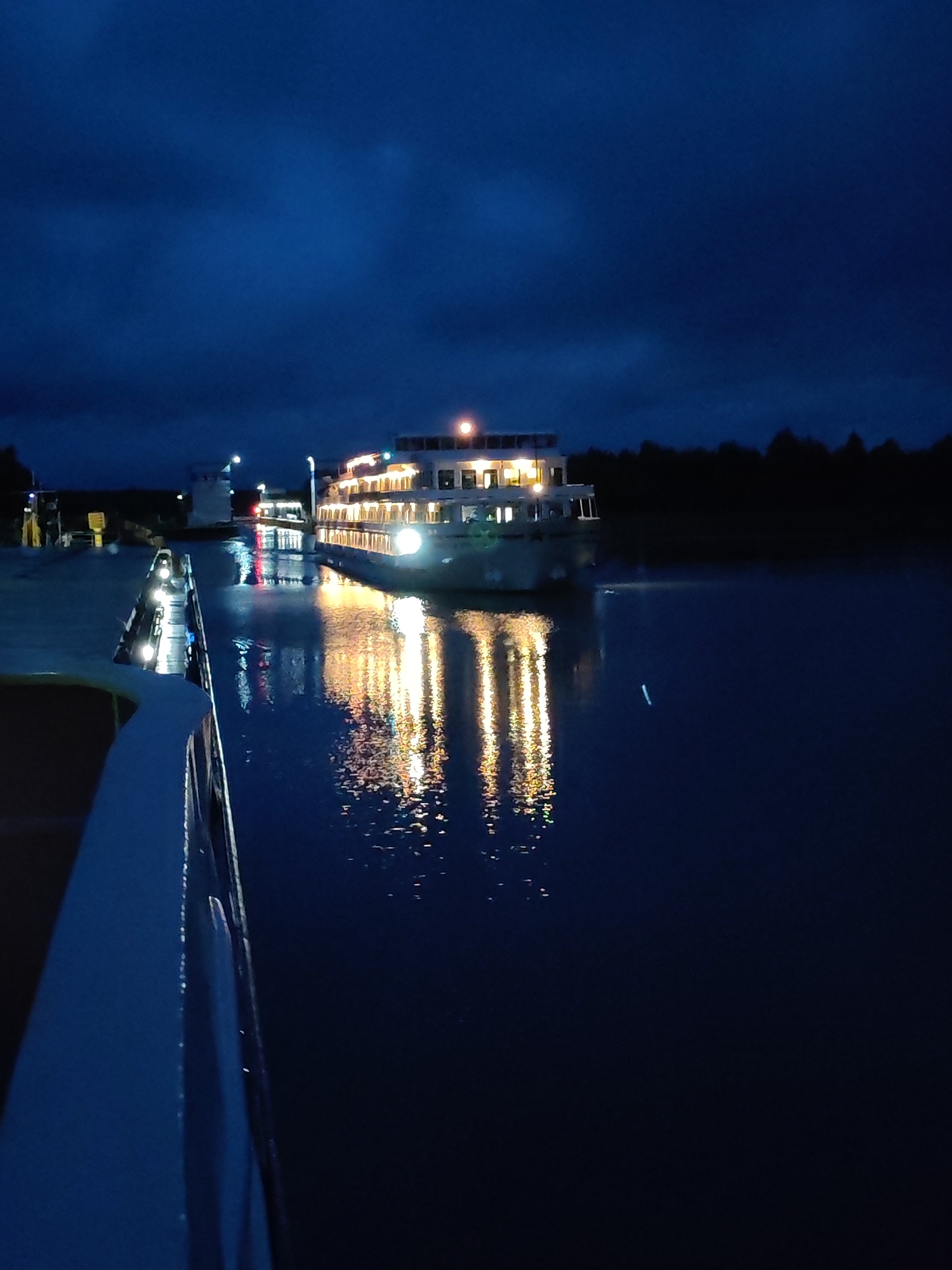 A night scene captures a large, multi-decked white ferry illuminated by numerous yellow lights, docked on a dark, calm body of water. The ferry's lights are reflected on the water's surface, creating a shimmering effect. In the foreground, the edge of another vessel, also white, is visible from an elevated perspective, extending from the bottom left of the frame towards the right. The background features a dark, silhouetted treeline against a deep blue, cloudy night sky. The location is identified as Devyatiny, Russia.