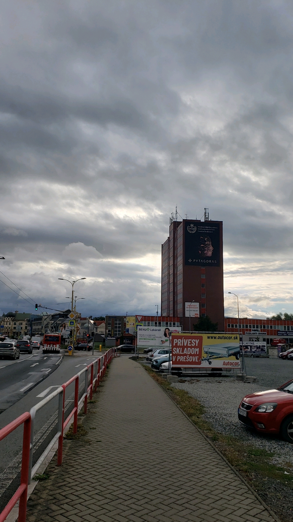 The image depicts a cloudy day in Prešov, Slovakia, focusing on a street scene with a tall, reddish-brown building dominating the background. The building has a large advertisement on its upper section featuring the text "PYTHAGORAS" and an image of a geometric shape. Adjacent to this building is a lower, beige-colored commercial structure with various signs, including "BENU Lekáreň" (BENU Pharmacy) and advertisements for "Autocar" and trailers for rent.
In the foreground, a paved pedestrian walkway runs alongside a street with traffic. Cars are visible on the road, moving in both directions. A red and white metal railing separates the walkway from the street. The sky is filled with heavy, gray clouds, suggesting overcast weather. The scene appears to be a commercial or business district within the city. There are no visible people in the immediate foreground.
