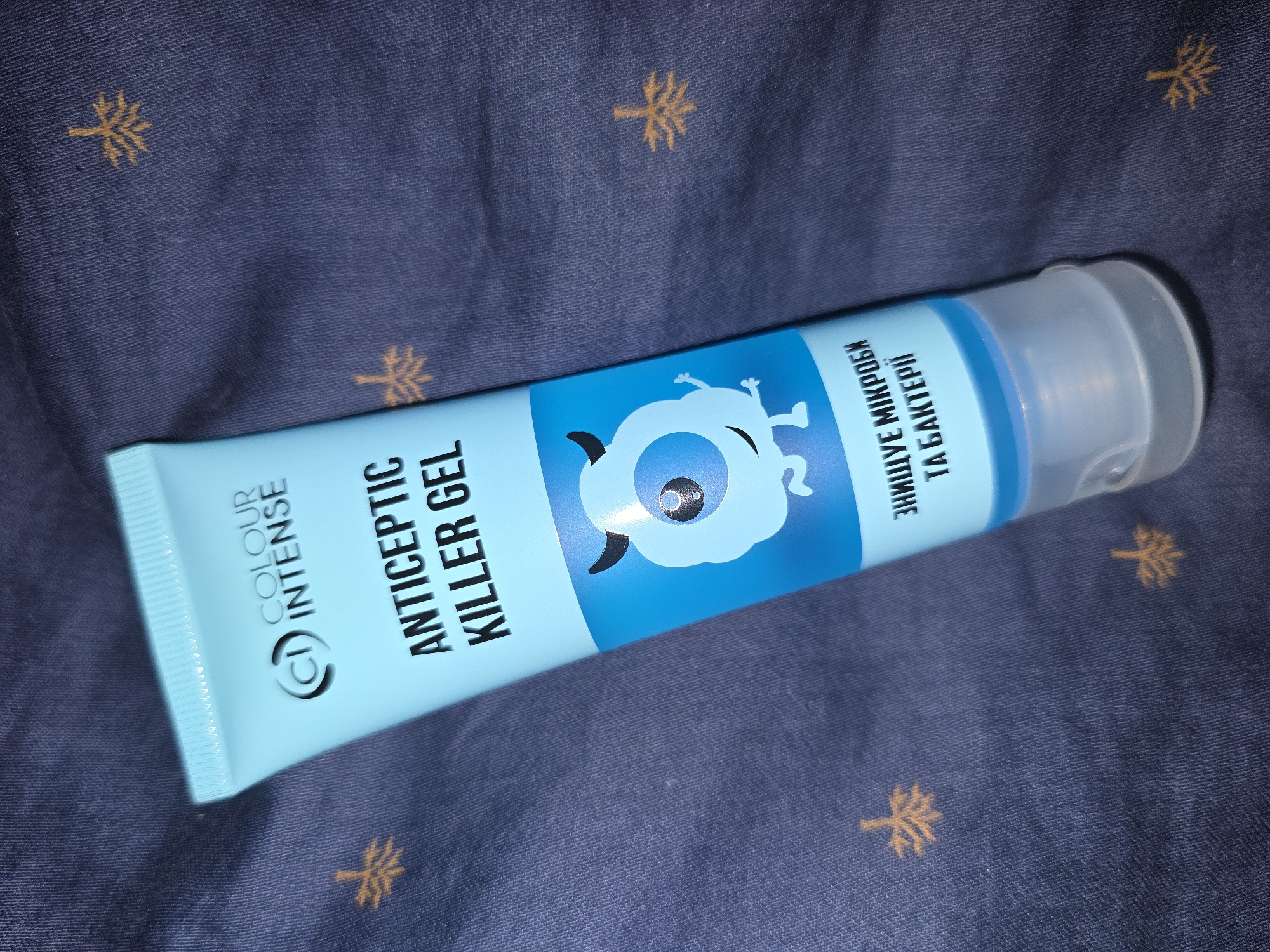The media file shows a tube of "Colour Intense" "Antiseptic Killer Gel" lying on a dark blue fabric with a pattern of small, stylized gold palm trees. The tube is light blue and has text printed on it in black and white. On the left side, the text reads "COLOUR INTENSE" and a logo that looks like a "CI" inside a circle. In the center, it says "ANTICEPTIC KILLER GEL" in large, bold, white letters. To the right of this, there is a cartoonish illustration of a blue, one-eyed monster with arms and legs. Further to the right, there is text in Ukrainian that reads "ЗНИЩУЄ МІКРОБИ ТА БАКТЕРІЇ" which translates to "DESTROYS MICROBES AND BACTERIA". The cap of the tube is clear and plastic. The lighting suggests the photo was taken indoors, possibly with a flash, creating some glare on the tube. The background fabric appears to be a type of textile, possibly cotton or a blend, with a slightly textured surface. The overall impression is that of a product photo, showcasing the antiseptic gel. The location context provided indicates Petropavlivska Borshchahivka, Ukraine, but there are no specific visual cues within the image to confirm this.