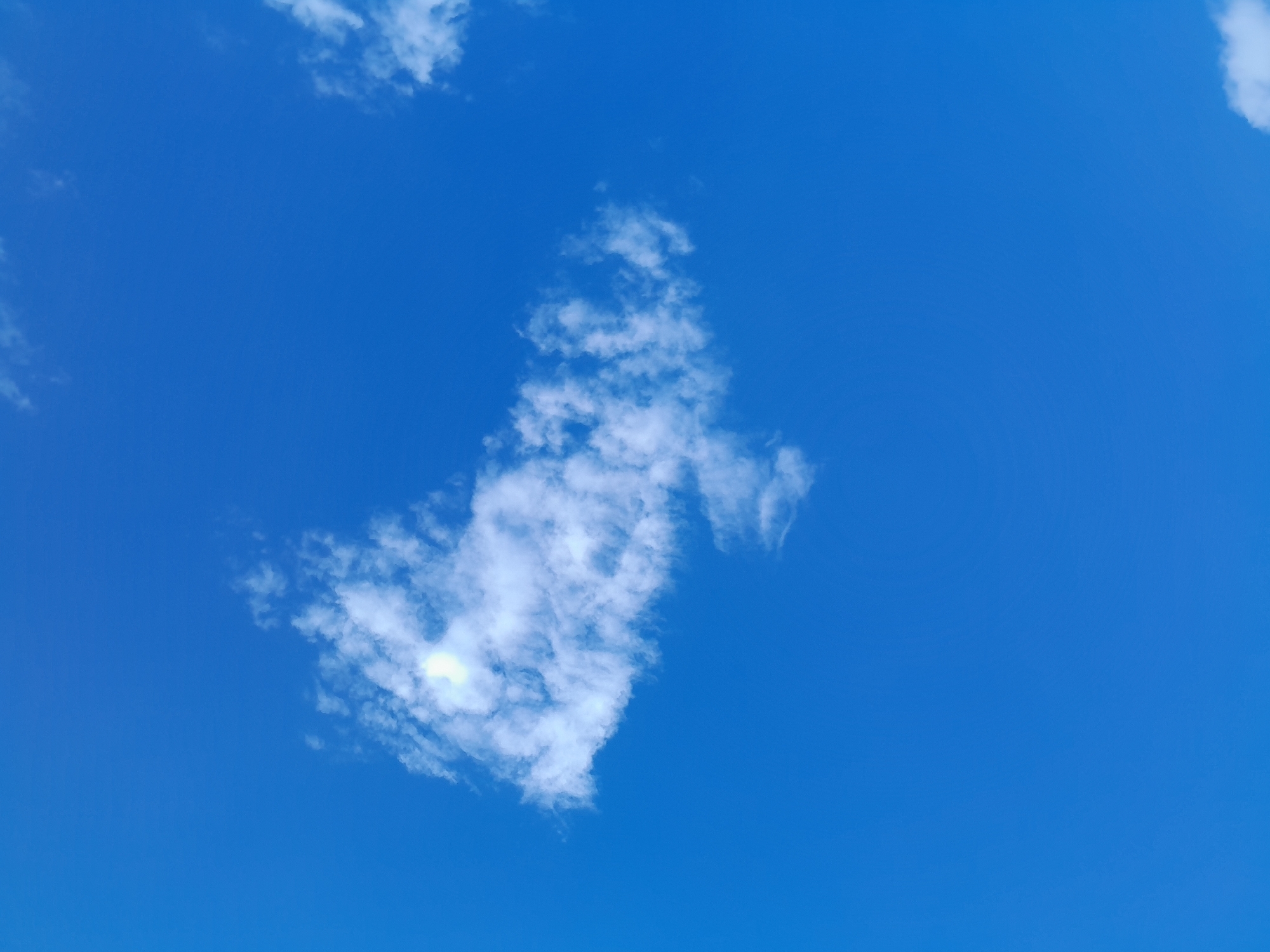 A bright blue sky is the background of this image, with a single, wispy cloud formation located slightly to the left of the center. The cloud is white and appears to be illuminated from behind by a bright light, possibly the sun, which is partially obscured by the cloud. The rest of the sky is clear, with no other significant features.