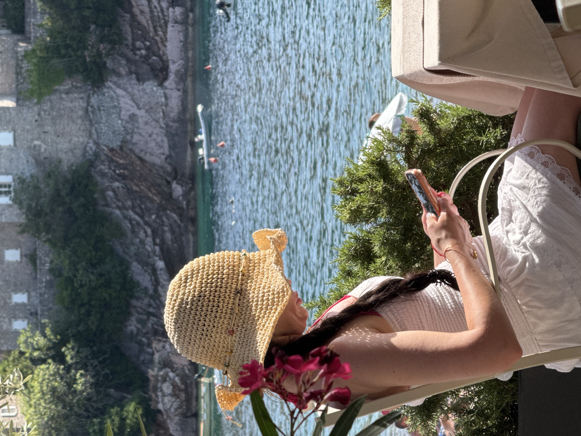 A woman is depicted seated at an outdoor table with a view of the Sveti Stefan coastline in Montenegro. She is positioned on the right half of the vertical frame, facing left with her head tilted down, engaged with a smartphone held in both hands. She wears a light-colored, possibly white or cream, strapless or sleeveless dress with a ruched bodice and a lacy hem, paired with a wide-brimmed straw hat adorned with small, multi-colored beads around the crown. Her long, dark hair is styled in a braid resting on her right shoulder. A delicate gold-toned bracelet is visible on her left wrist.
In the immediate foreground to her left, vibrant dark pink flowers and green foliage partially obscure her. To her right, a light-colored tablecloth covers a table. Directly behind her and the table, a dense green shrub, likely rosemary, is visible in a dark planter.
The midground features a body of calm, blue-green water with subtle ripples, characteristic of the Adriatic Sea. Several small watercraft are present: a white inflatable boat with an outboard motor floats on the left, accompanied by several orange buoys. Another darker boat is partially visible on the far right.
The background showcases the distinctive fortified stone buildings of Sveti Stefan, with their gray facades and small, rectangular windows, nestled amidst green trees and shrubs on a rocky promontory. The scene is brightly lit by natural sunlight, casting shadows and creating dappled light effects on the woman's arm.