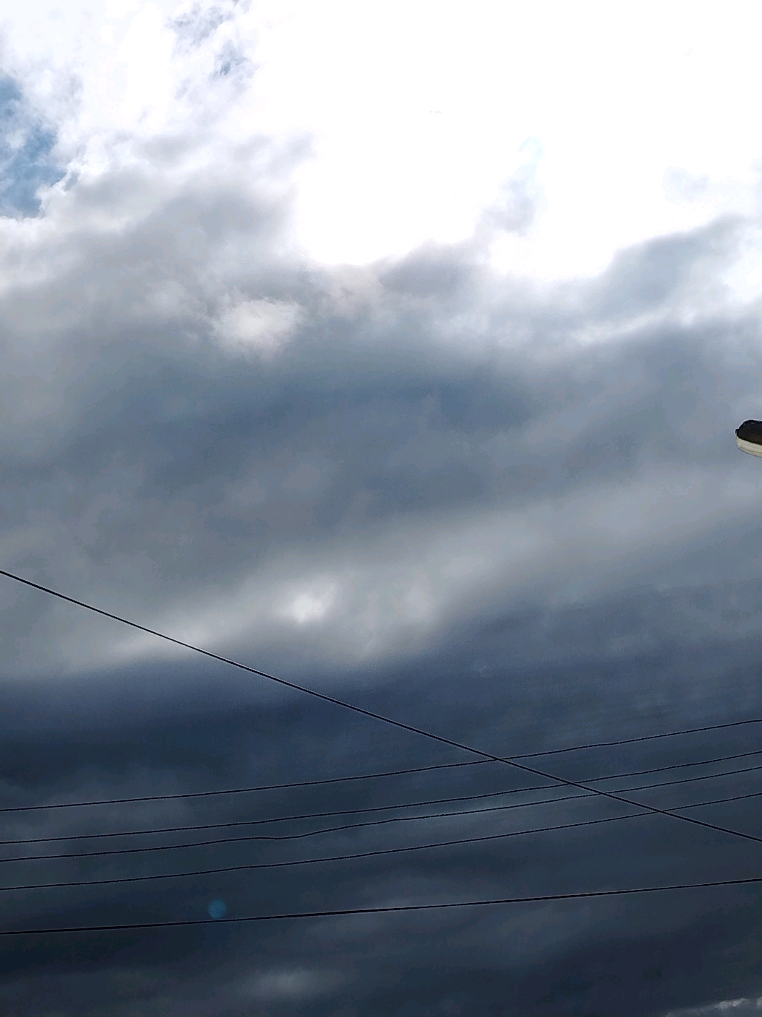 The image shows a cloudy sky with power lines crisscrossing the frame. In the upper right corner, part of a street lamp is visible. The sky transitions from bright white clouds at the top to dark, ominous clouds lower down. The overall scene suggests an overcast day, possibly before or during a storm. The location is identified as Muravlenko, Russia.