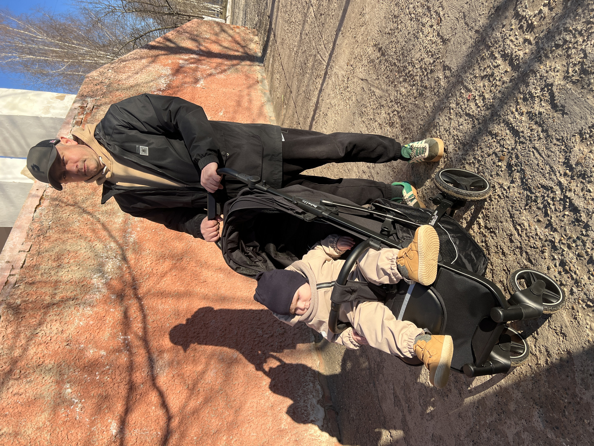 A man is standing next to a baby in a stroller. The man is wearing a black jacket, black pants, and green and brown sneakers. He has a black cap on. The baby is wearing a beige jacket, pants, and tan shoes. The stroller is black. The location appears to be an outdoor area with concrete and a painted wall. The image may have been taken in Noviy, Russia. The image is rotated clockwise in post-processing.