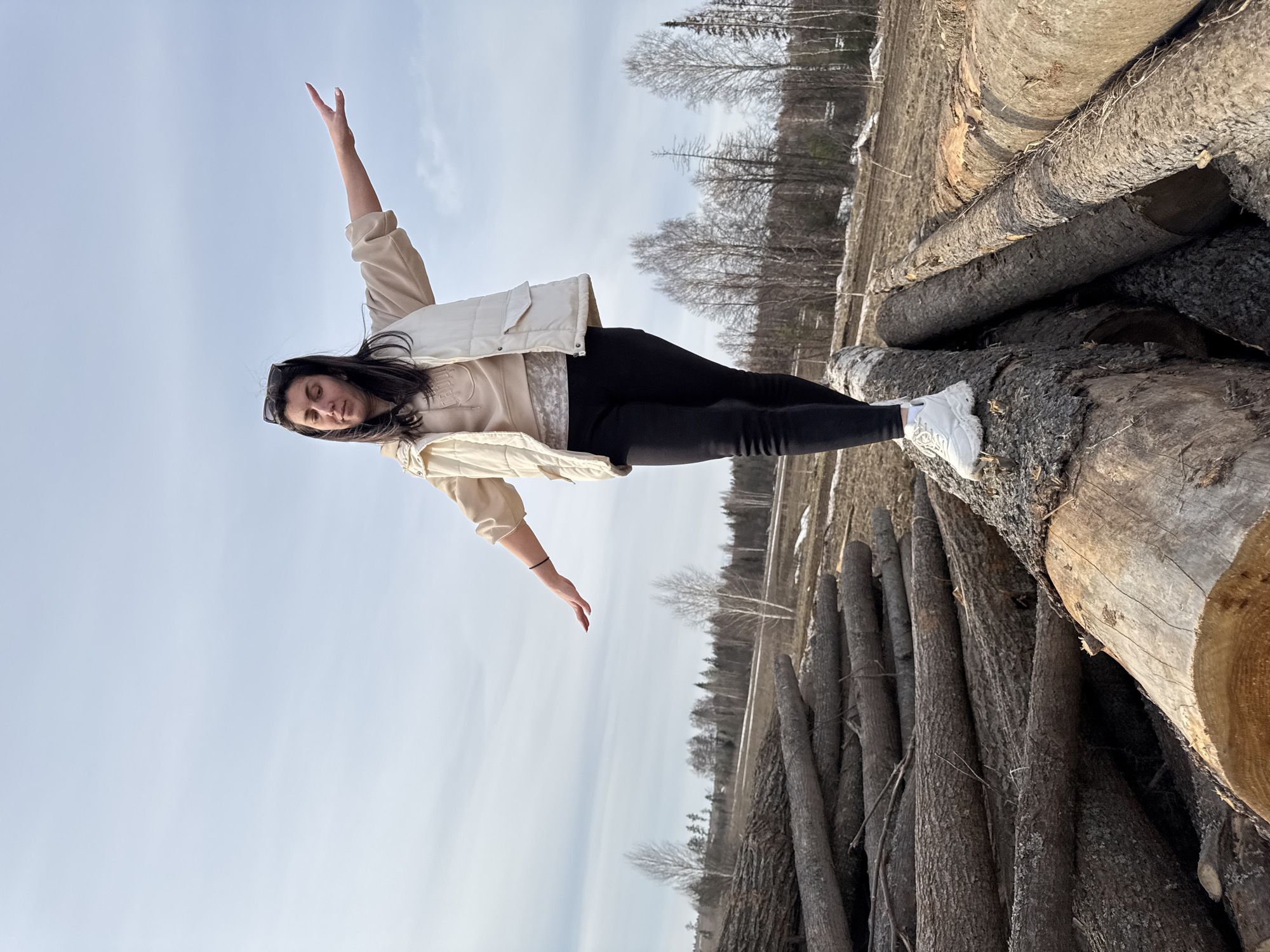 The image depicts a young woman lying atop a stack of logs. She is wearing black leggings, a beige sweatshirt and a white puffer vest. Her arms are outstretched, one pointing towards the sky and the other towards the horizon. The background features a clear sky and a row of trees in the distance. The setting is a lumber yard in Van'ki, Russia.