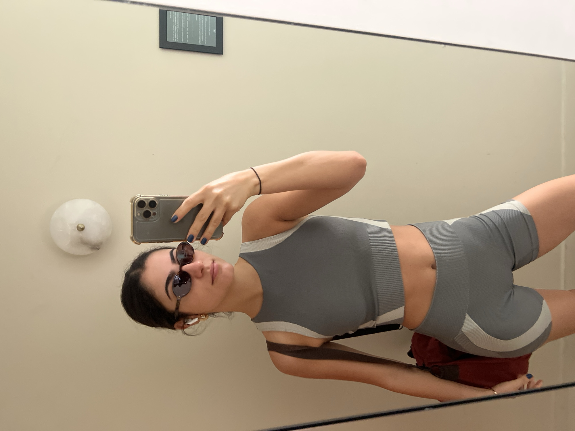 This media file shows a woman taking a selfie in a mirror. She is wearing a grey athletic outfit consisting of a crop top and shorts. She has dark hair pulled back and is wearing sunglasses and white wireless earbuds. Her left hand is holding a smartphone, and her right arm is bent at the elbow. The background appears to be a wall with a light fixture on the left and a framed object hanging on the wall in the upper left quadrant. The image is taken indoors, likely in a fitting room or dressing area, given the mirror and the clean, neutral walls. The lighting is soft and even, suggesting it could be daytime or a well-lit interior. There is no discernible weather information, but the setting implies an indoor environment. The overall mood seems casual and possibly a moment of self-appreciation or outfit checking. The woman's expression is neutral, focusing on the selfie. No specific text is legible within the image, except for what might be on the framed object in the background, which is too blurry to read. The scene does not contain any obvious Parisian landmarks or context.