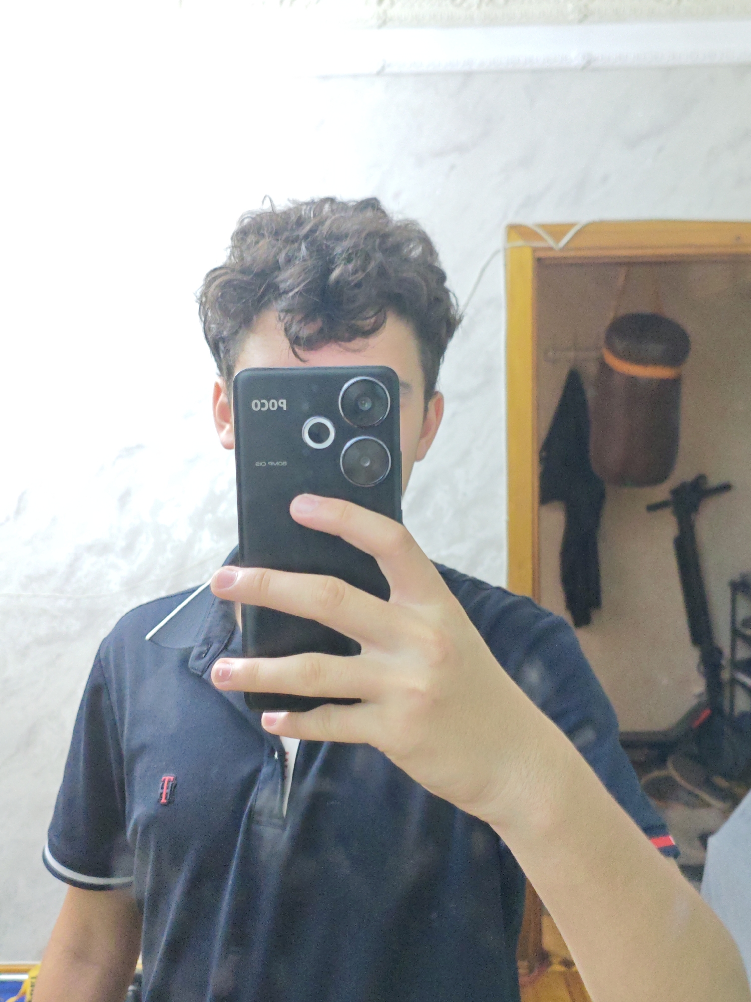 A young person is taking a mirror selfie in what appears to be a bathroom. They are holding a black smartphone in their right hand, obscuring their face. The phone displays the "POCO" logo and other text that is reversed due to the mirror. The person is wearing a dark blue polo shirt with white trim on the collar and sleeves. A small red embroidered logo, possibly an initial, is visible on the left side of the shirt.
The background shows a tiled wall to the left, and a mirror frame to the right, with a yellow border. Reflected in the mirror are various objects, including what looks like a dark brown container with an orange band, a black kitchen knife, and a black electric scooter. The lighting suggests it could be daytime, but the exact time is not determinable. The setting appears to be domestic, likely a home bathroom. The person's expression cannot be discerned due to the phone covering their face, but the act of taking a selfie implies a moment of self-documentation or casual sharing. The overall mood is informal and everyday. The location context of Baku City, Azerbaijan, provides a geographical setting for this personal snapshot.