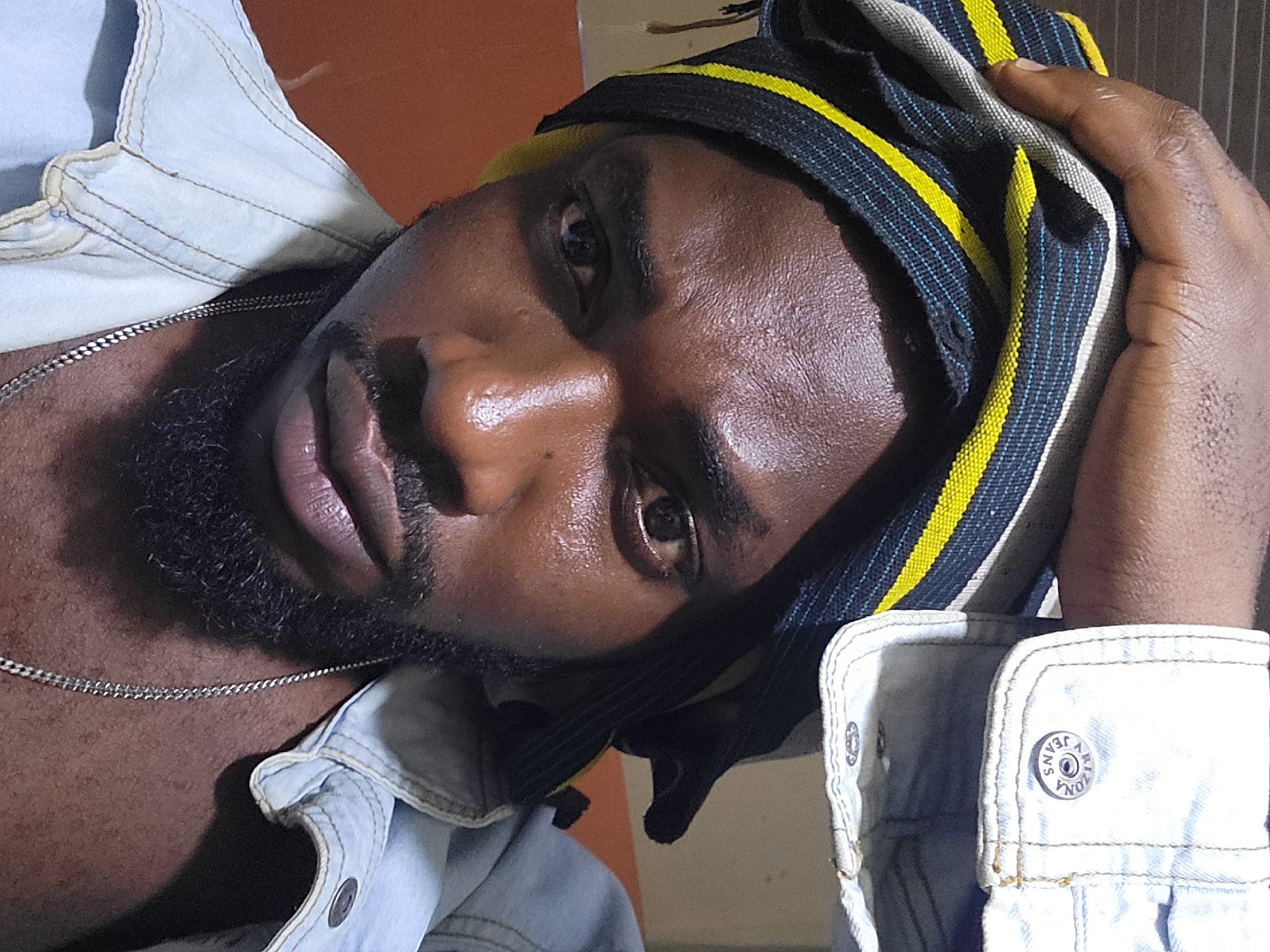 A close-up, low-angle shot of a dark-skinned man with a beard, partially covered by a black, yellow, and white striped head covering. He is wearing a light blue denim shirt and a silver chain. The background is blurred and indistinct but appears to be an interior setting, however, the specific location in Jos, Nigeria cannot be determined.