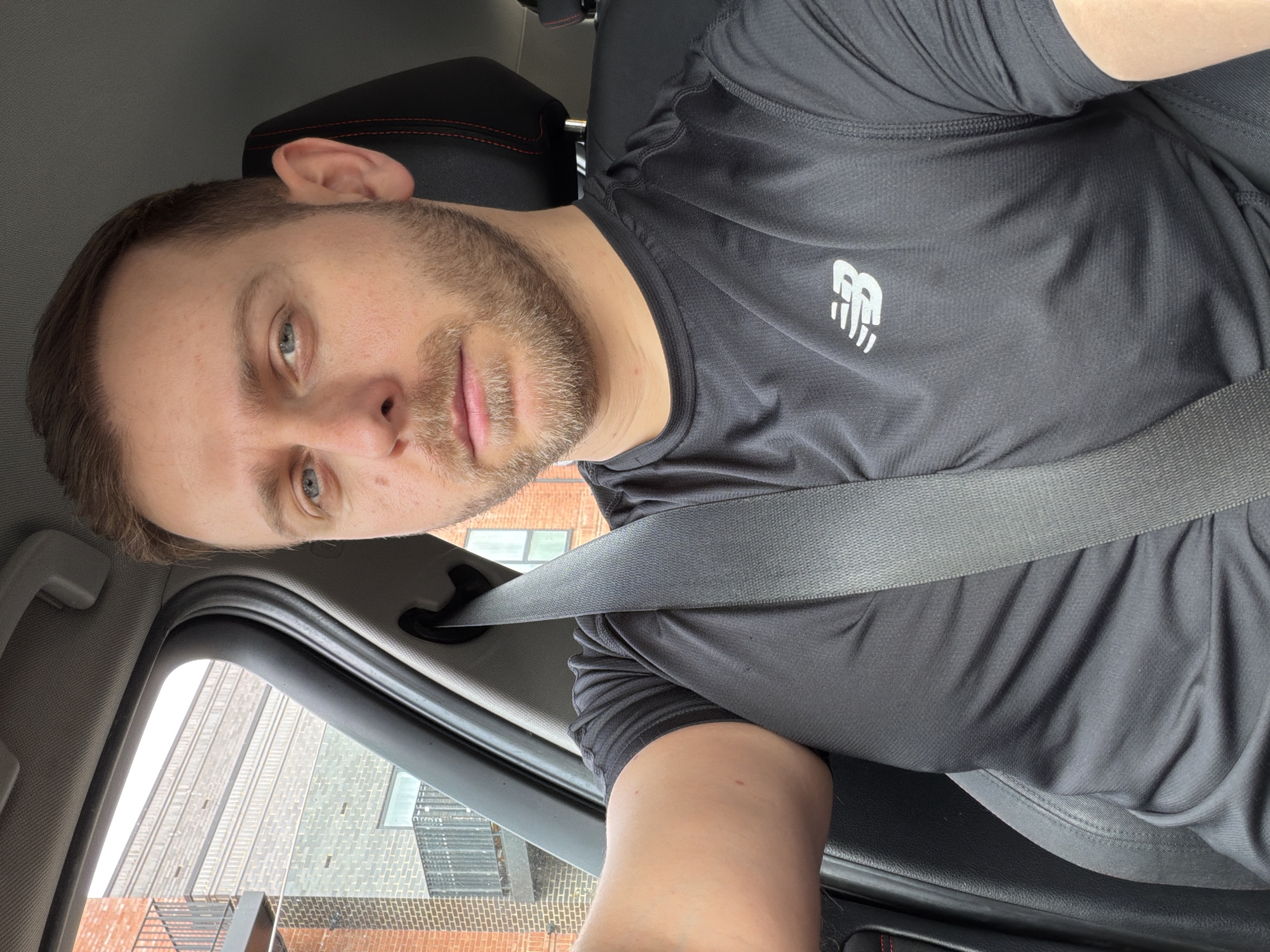 A man is wearing a black New Balance t-shirt and a seatbelt in a car. He has short brown hair, blue eyes, and light stubble. Outside the car window is a view of brick buildings, likely residential or commercial. There is no definitive information to confirm the location is London, United Kingdom.