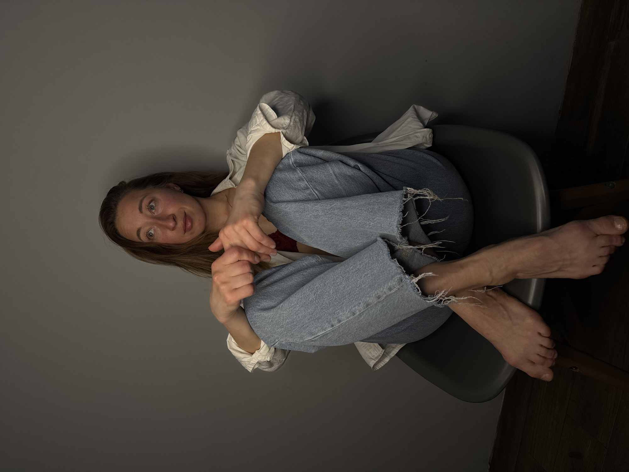 A young woman with long brown hair is sitting in a gray chair against a gray wall in a room with a dark wooden floor. She is wearing blue jeans and a white shirt, with a hint of red clothing visible underneath. She is barefoot, and her arms are crossed in front of her. The location is Chaikovsky, Russia.