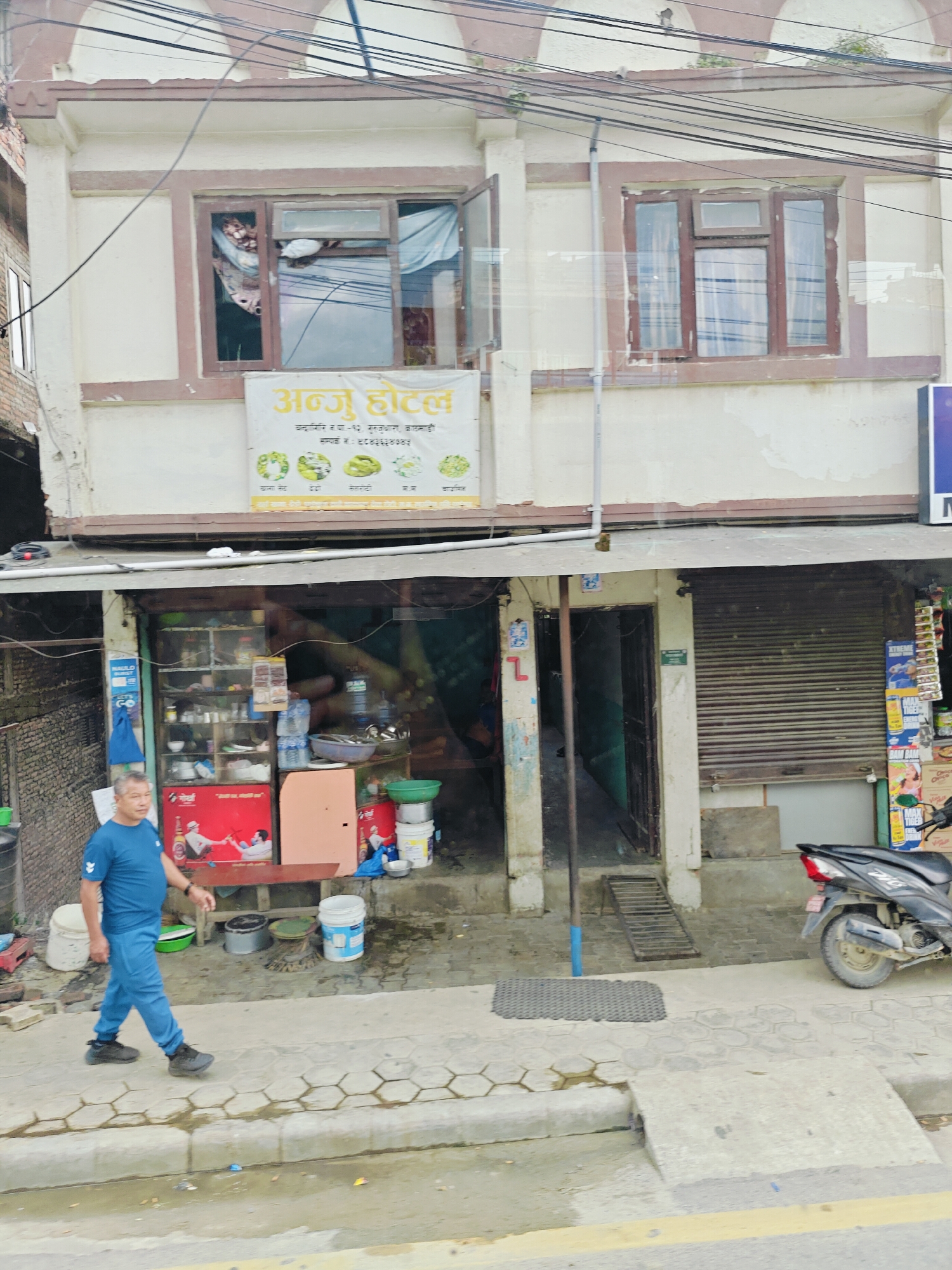 The image depicts a street scene in Chandragiri, Nepal. A man in a blue t-shirt and blue pants walks on a sidewalk in front of a building. The building has a signage that reads "अन्नु होटल" (Annu Hotel) in Nepali script. Below the signage, there are several small circular images of food items. The ground floor of the building appears to be a shop or restaurant, with shelves visible through an open doorway, displaying various items including bottles and containers. A black motorcycle is parked on the right side of the image. The environment appears to be an urban setting with overhead electrical wires. The sidewalk is paved with hexagonal tiles.