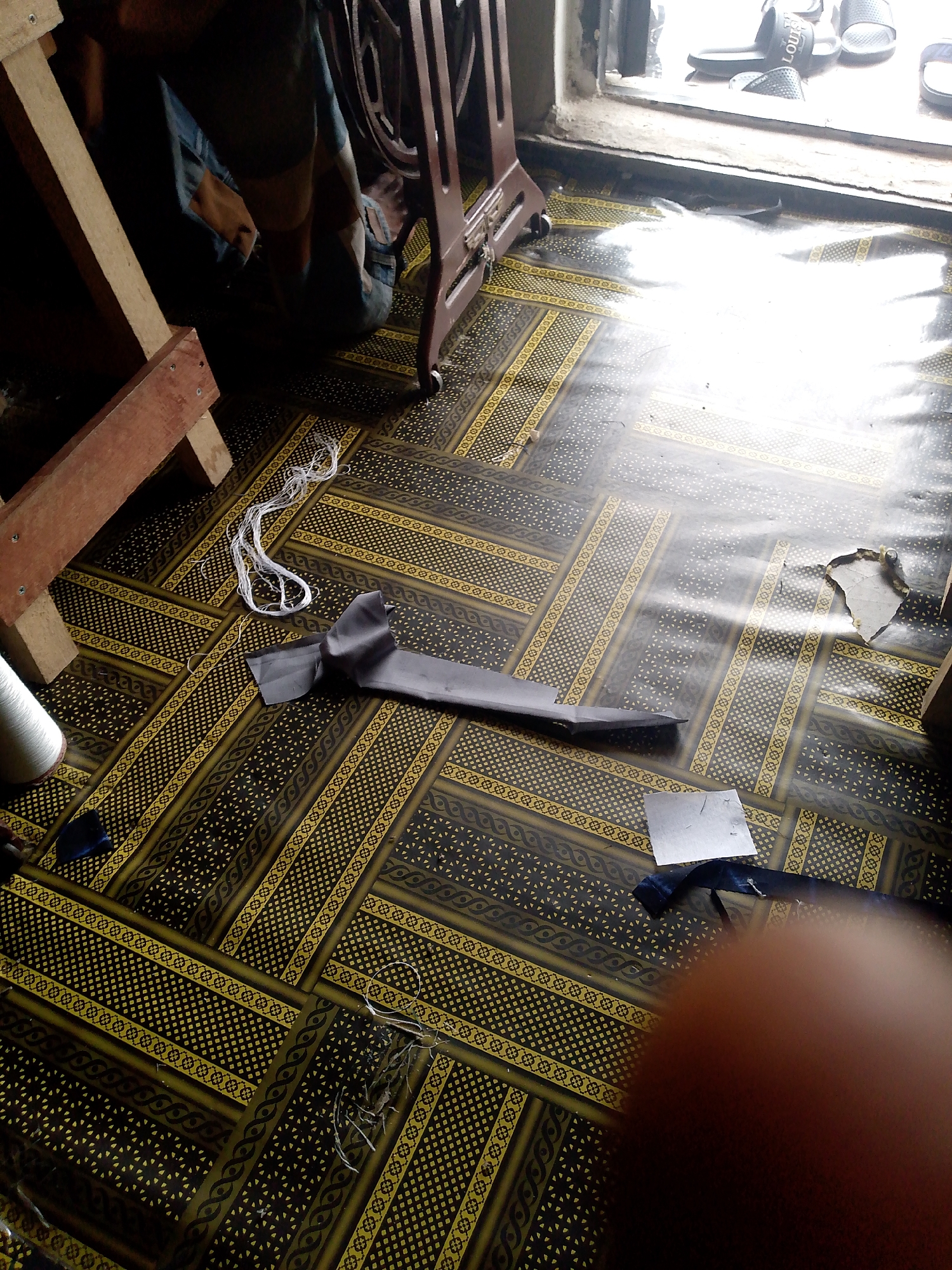 The image depicts an indoor scene with a person, likely engaged in tailoring, in the city of Jauro Yinu, Nigeria. The floor is covered in a patterned linoleum. A vintage sewing machine is partially visible on the right side of the frame, with a wooden frame structure on the left. Scattered on the floor are various sewing materials, including a spool of white thread, a piece of dark fabric folded over, a small square of paper, and loose white threads. In the background, a window shows a bright outdoor light, with several pairs of slippers visible on the windowsill. The person's legs, clad in blue jeans, are bent as if working on the floor. The overall impression is of a functional workspace where sewing activities are taking place.