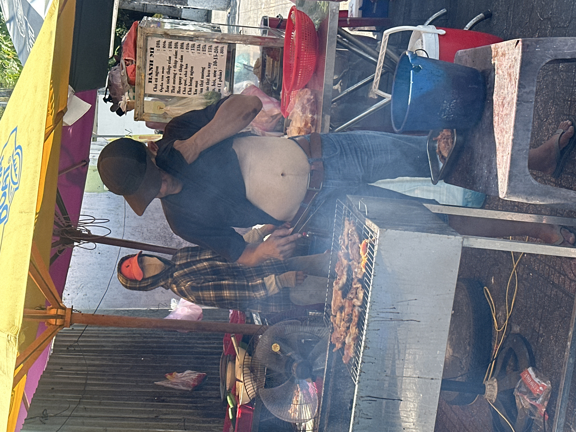 Captured in Nha Trang Ward, Vietnam, the scene depicts two men at a streetside food stall under a yellow umbrella displaying "Bánh Mì" offerings. One man, with a visible belly, is dressed in jeans and a dark top, wearing a baseball cap, seemingly tending to the stall. A second person, in a plaid jacket and red cap, is also present.
The stall showcases a grill with cooking meat, and a menu board listing various "Bánh Mì" options. A fan is positioned nearby, likely for ventilation. Adjacent to the stall are miscellaneous items, including buckets, a red basket, and a white stool.