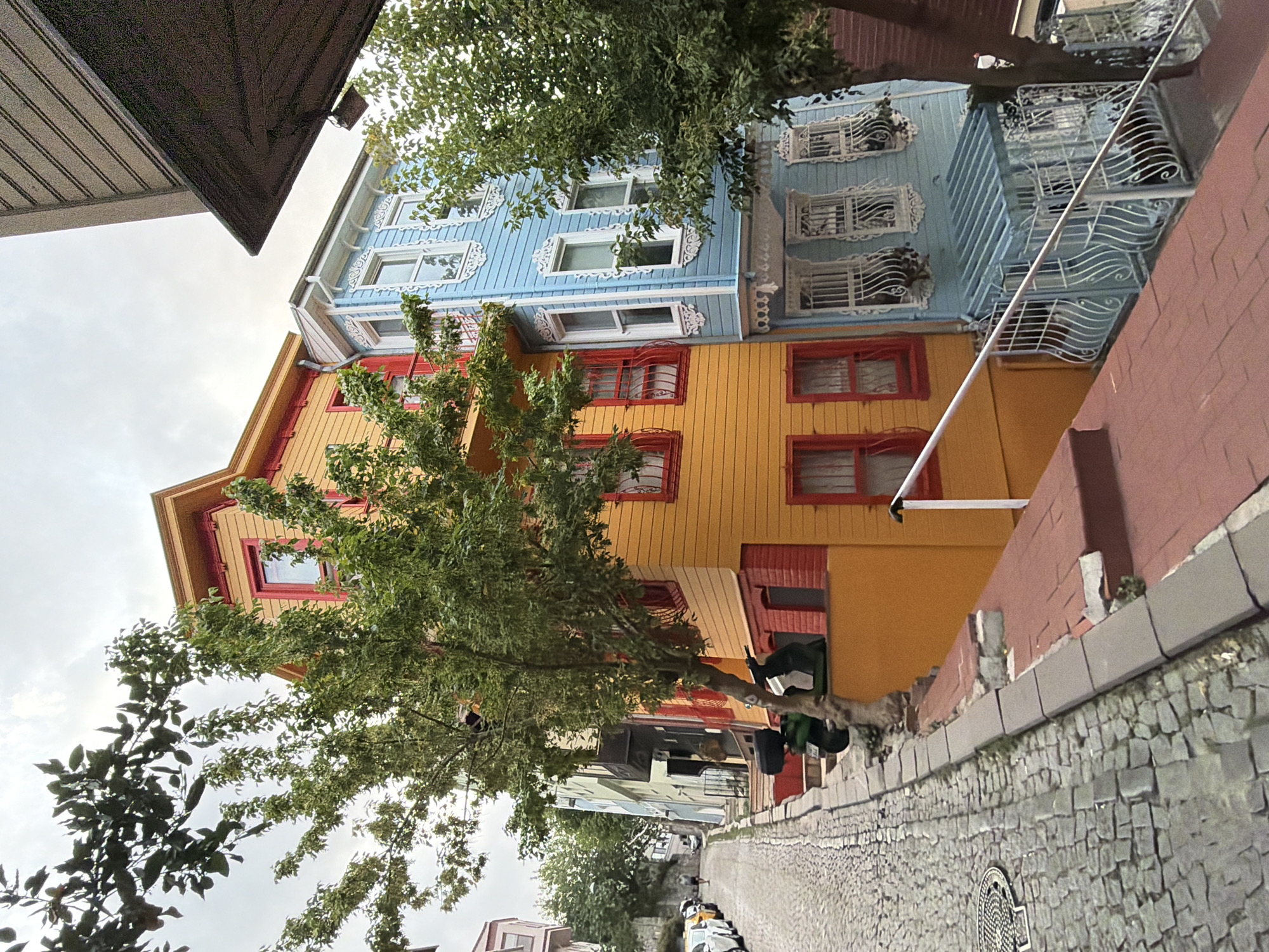 The image depicts a street scene in Üsküdar, Turkey, characterized by colorful, traditional Ottoman-style houses lining a cobblestone street that slopes upwards. On the left, a row of parked cars, including white sedans and a yellow taxi, are visible on the lower section of the street. Further up the street, a few pedestrians are walking.
The architecture of the houses is a prominent feature. The house closest to the foreground on the right is painted a vibrant orange with red trim around the windows and on the lower brick section. A large green tree with dense foliage grows in front of this house, partially obscuring its facade. Adjacent to the orange house is a light blue house with intricate white decorative woodwork around its windows and a bay window structure on the lower level, also featuring white ornamental metalwork. Another tree with greenery is situated to the right of the blue house.
The cobblestone street is bordered by a raised curb made of concrete blocks. The street shows signs of wear and some repair work on the brick steps leading up to the houses. The sky is overcast, suggesting a cloudy day. The overall impression is a charming, historic urban environment.