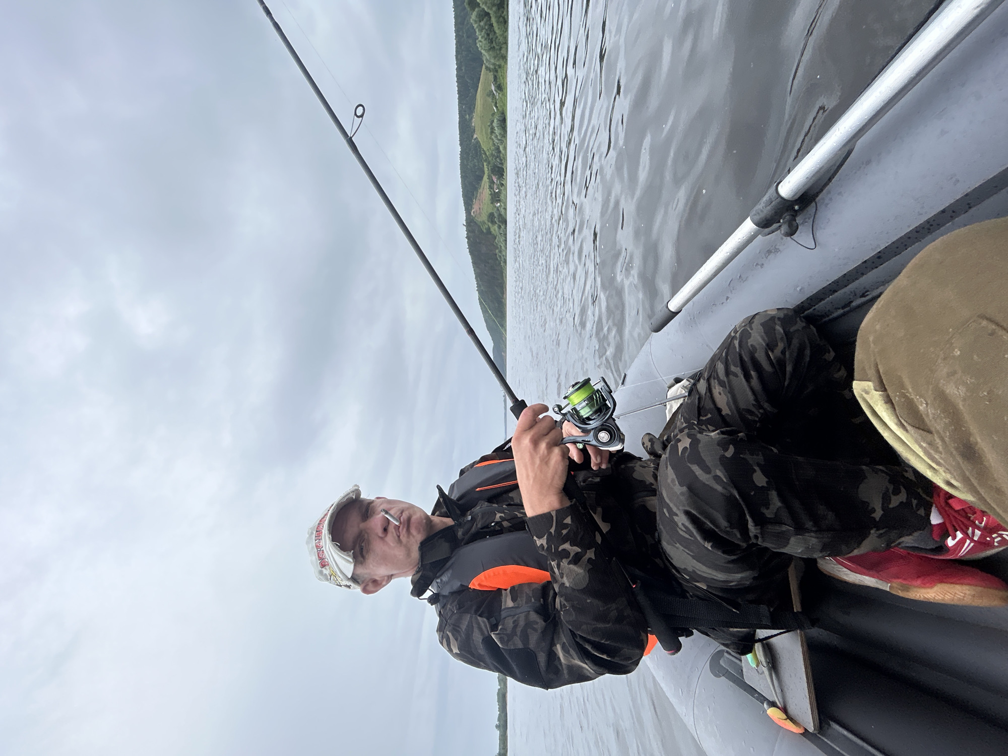 A man is fishing from an inflatable boat on a river. He is wearing a camouflage jacket, a life vest, and a baseball cap. He is holding a fishing rod and has a cigarette in his mouth. The boat is on a wide river with hills covered in trees in the background. The sky is overcast. The location is the city of Olkhovka, Russia.