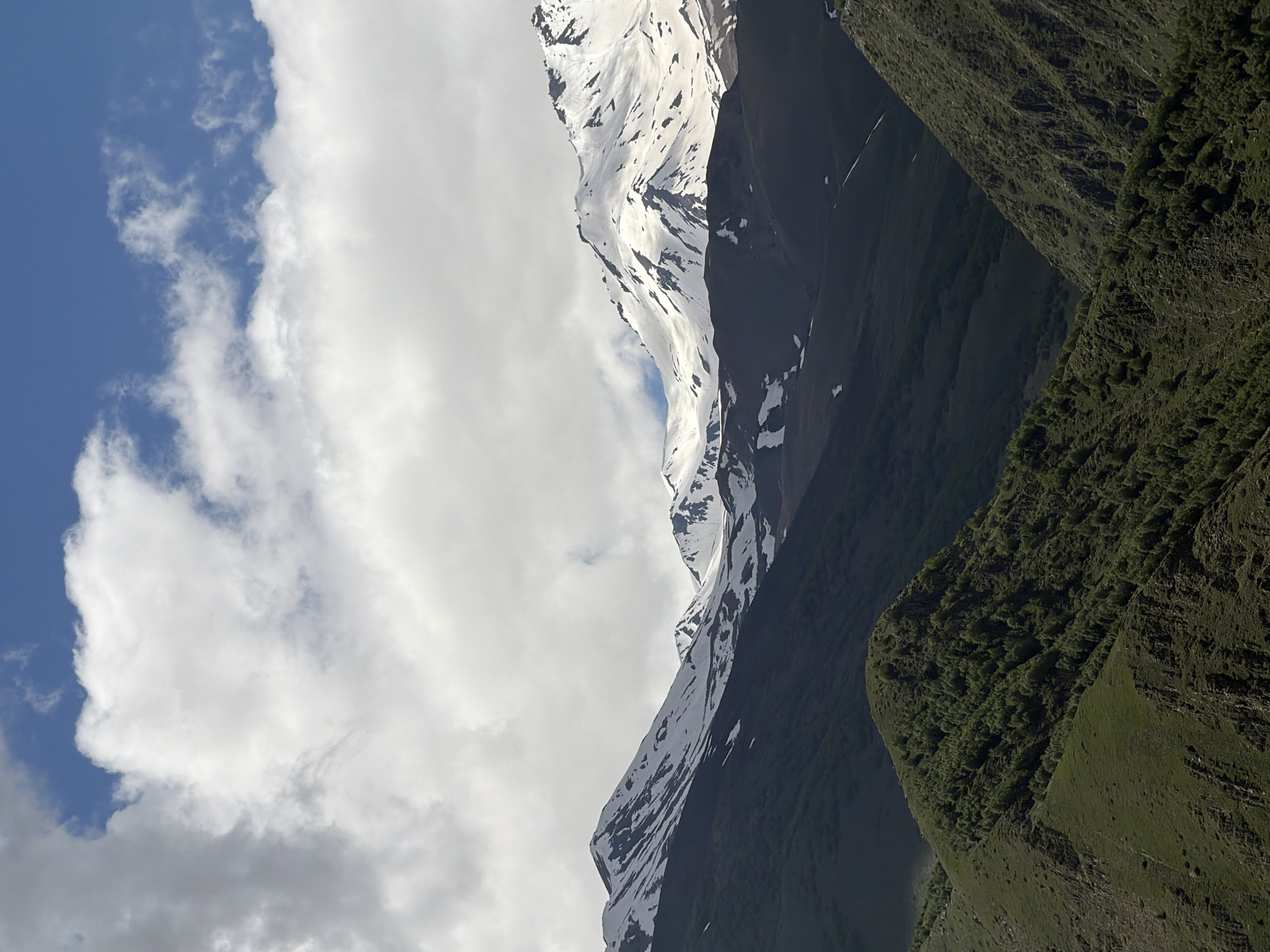 The image depicts a mountainous landscape with a sky filled with clouds. The scene is dominated by the imposing presence of snow-capped mountains. The snow covers the upper portions of the mountains, contrasting sharply with the darker, rocky slopes below. Patches of snow are scattered across the mountainsides, suggesting a rugged and untamed terrain.
The lower slopes of the mountains are covered in lush green vegetation, indicative of a lower altitude or a milder climate. The texture of the vegetation suggests a dense growth of trees or shrubs, creating a vibrant green carpet on the hillsides. The contrast between the snow-covered peaks and the green slopes creates a visually striking image of a mountain range.
The sky is a mix of blue and white. Large, puffy clouds are scattered across the sky, with some areas of clear blue visible. The clouds appear to be illuminated by sunlight, suggesting it is daytime. The lighting conditions create a dramatic interplay of light and shadow on the mountains, highlighting their contours and textures.
The orientation of the image is such that the landscape appears to be viewed from a slightly elevated position, looking upwards towards the peaks. The composition draws the viewer's eye from the green slopes at the bottom to the snow-capped summits and the expansive sky above. There are no people or man-made objects visible in the image, emphasizing the natural and wild character of the scene. The overall impression is one of grandeur and natural beauty.