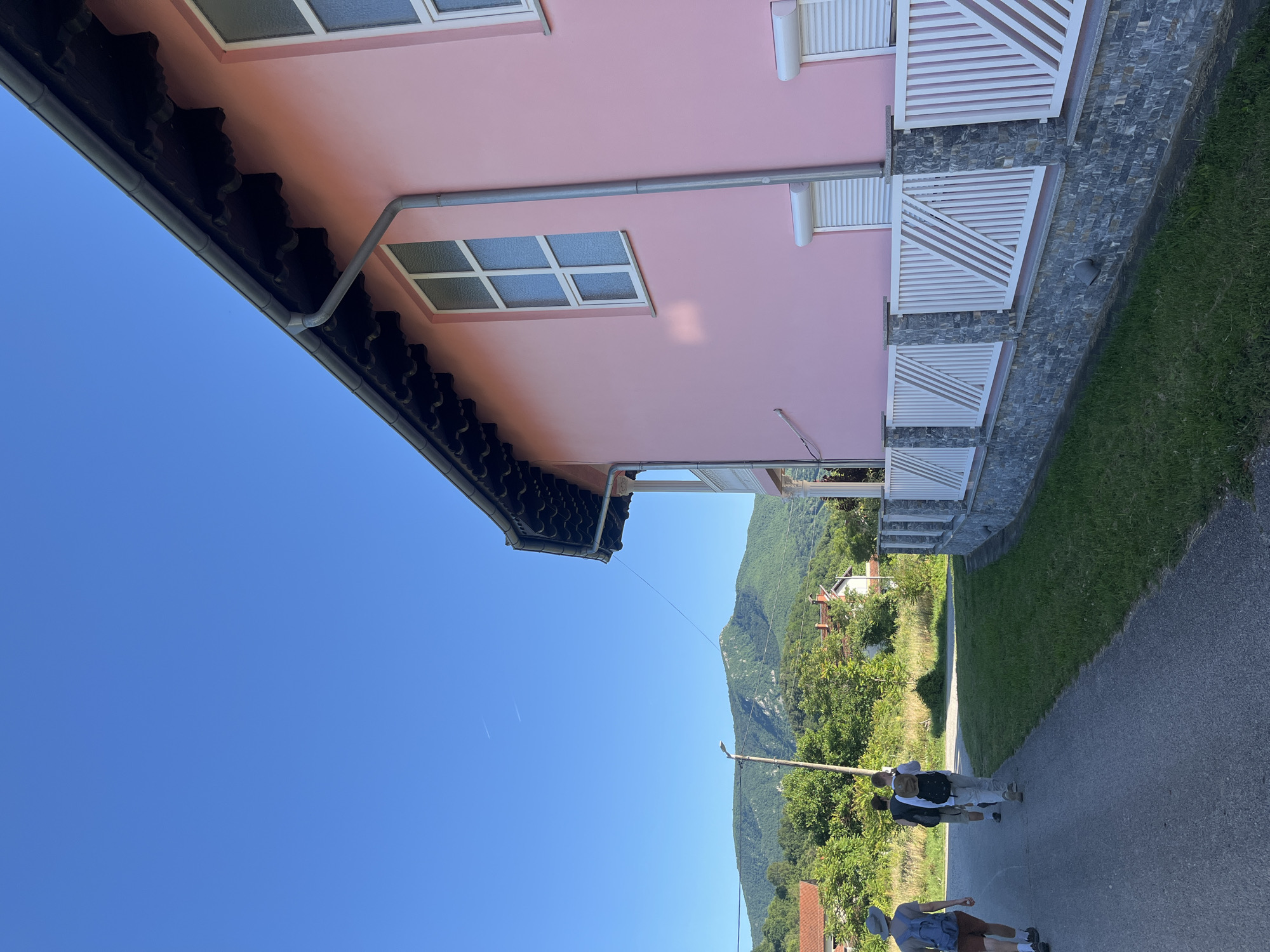 In Petrovac-na-Mlavi Municipality, Serbia, on a clear, sunny day, three individuals are walking away from the camera down a paved road. The person in the lead wears a hat and a blue backpack, with brown shorts and white socks. Behind them, a second individual carries a dark backpack, and the third person also has a backpack. They are walking alongside a property with a vibrant pink house. The house features white-framed windows, some with roller shutters. A white fence, supported by stone pillars and a stone base, separates the property from the road. Beyond the road and fence, a grassy area leads to a backdrop of lush green trees and a prominent, tree-covered mountain range under a bright blue sky, with two faint contrails visible.