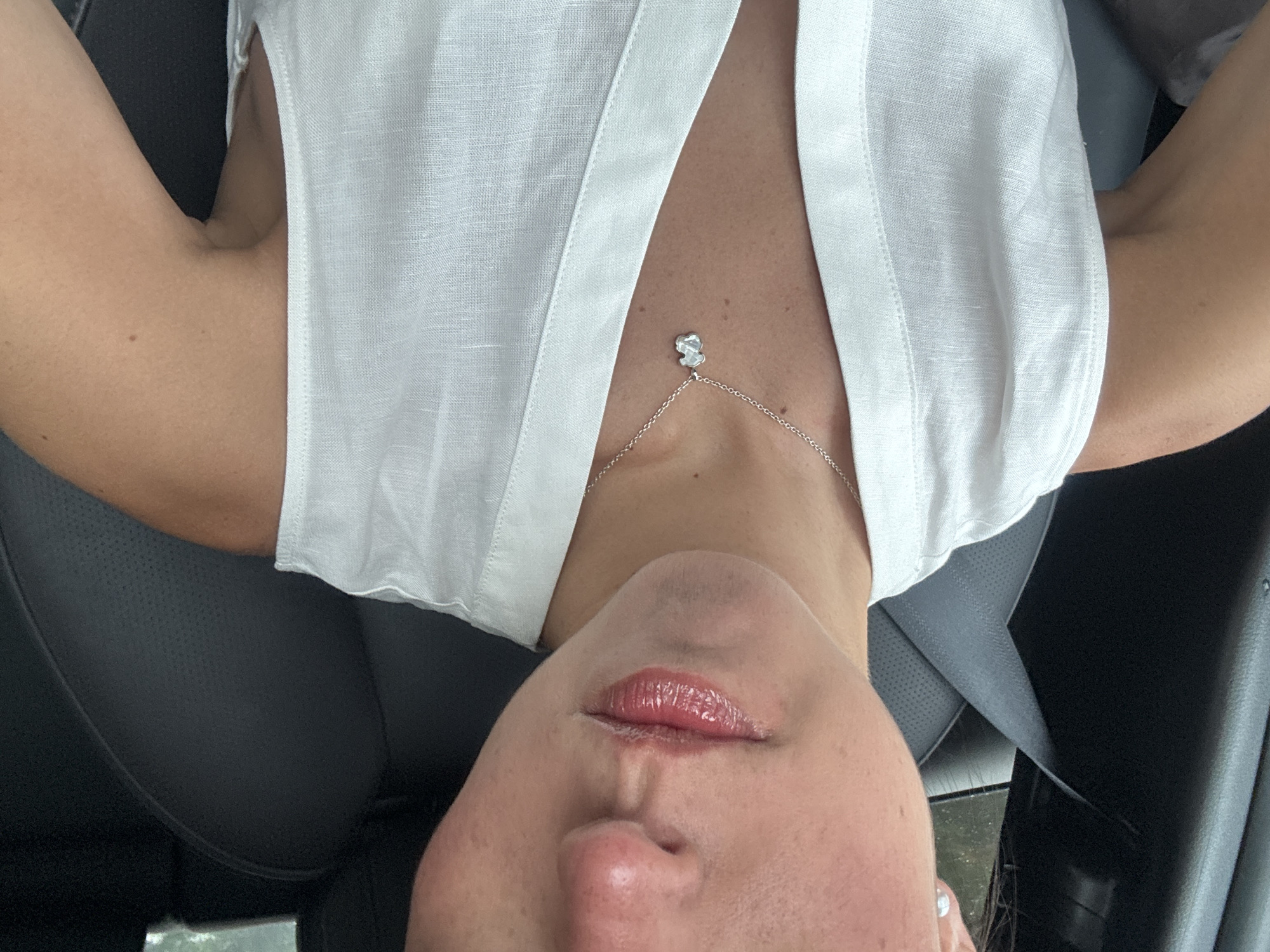 A person is shown from the chest up. The person is wearing a white shirt and a necklace with a pendant. The person is inside a vehicle. The location is in the city of Unknown, Unknown.