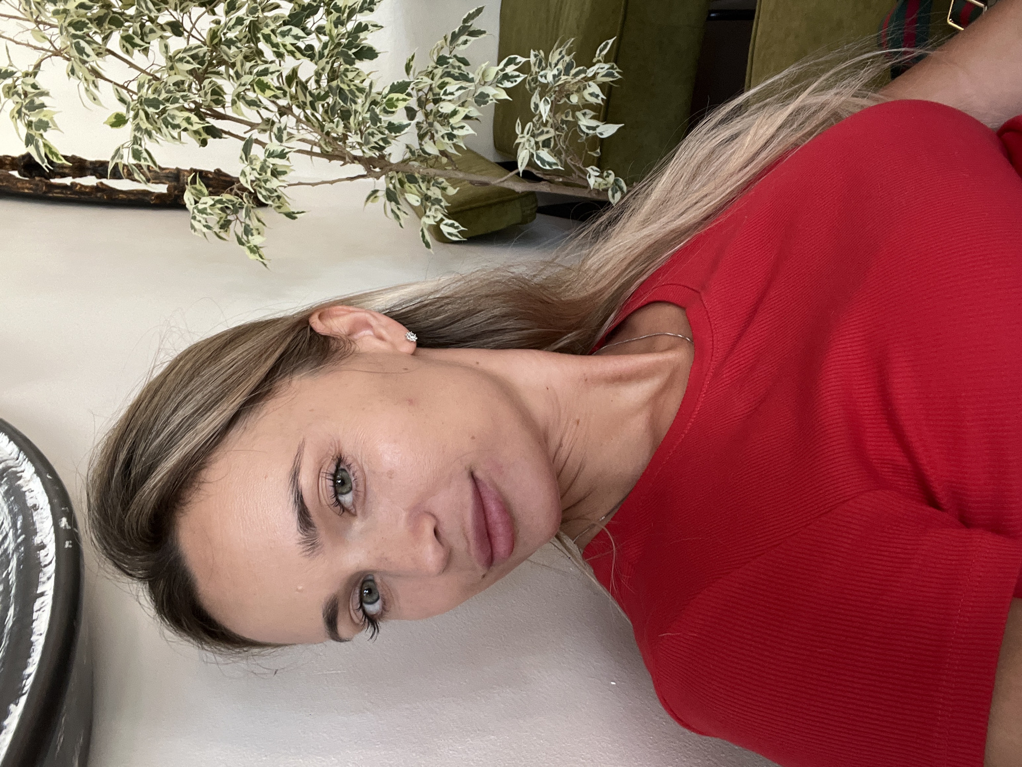 The image shows a young woman with blonde hair and green eyes. She is wearing a red top and a silver necklace. She is leaning against a white wall, and behind her is a plant and a green couch. The woman is looking at the camera and smiling slightly. The background appears to be an indoor setting, possibly a cafe or a living room. There is no indication of the specific location in Odesa, Ukraine.
