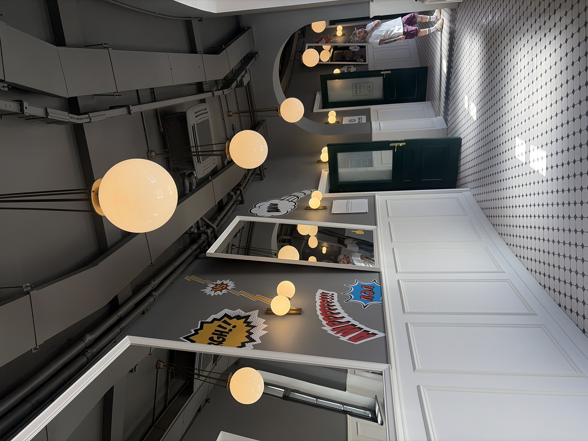 The image depicts an interior hallway of a establishment in Saint Petersburg, Russia. The hallway is lined with white paneled walls and a patterned tiled floor. Several large, round, illuminated light fixtures hang from the ceiling, which is exposed with visible ductwork and pipes. The walls are decorated with comic-book-style graphics.
On the left side of the hallway, a large mirror reflects the opposite wall, showing more of the comic-style art and ceiling lights. Further down the hallway, a man wearing a white t-shirt and purple shorts stands near two dark green doors. One door is slightly ajar, revealing a glimpse of another room. The sunlight casts shadows across the tiled floor, indicating natural light entering from an unseen source. The overall aesthetic suggests a modern and artistic design.