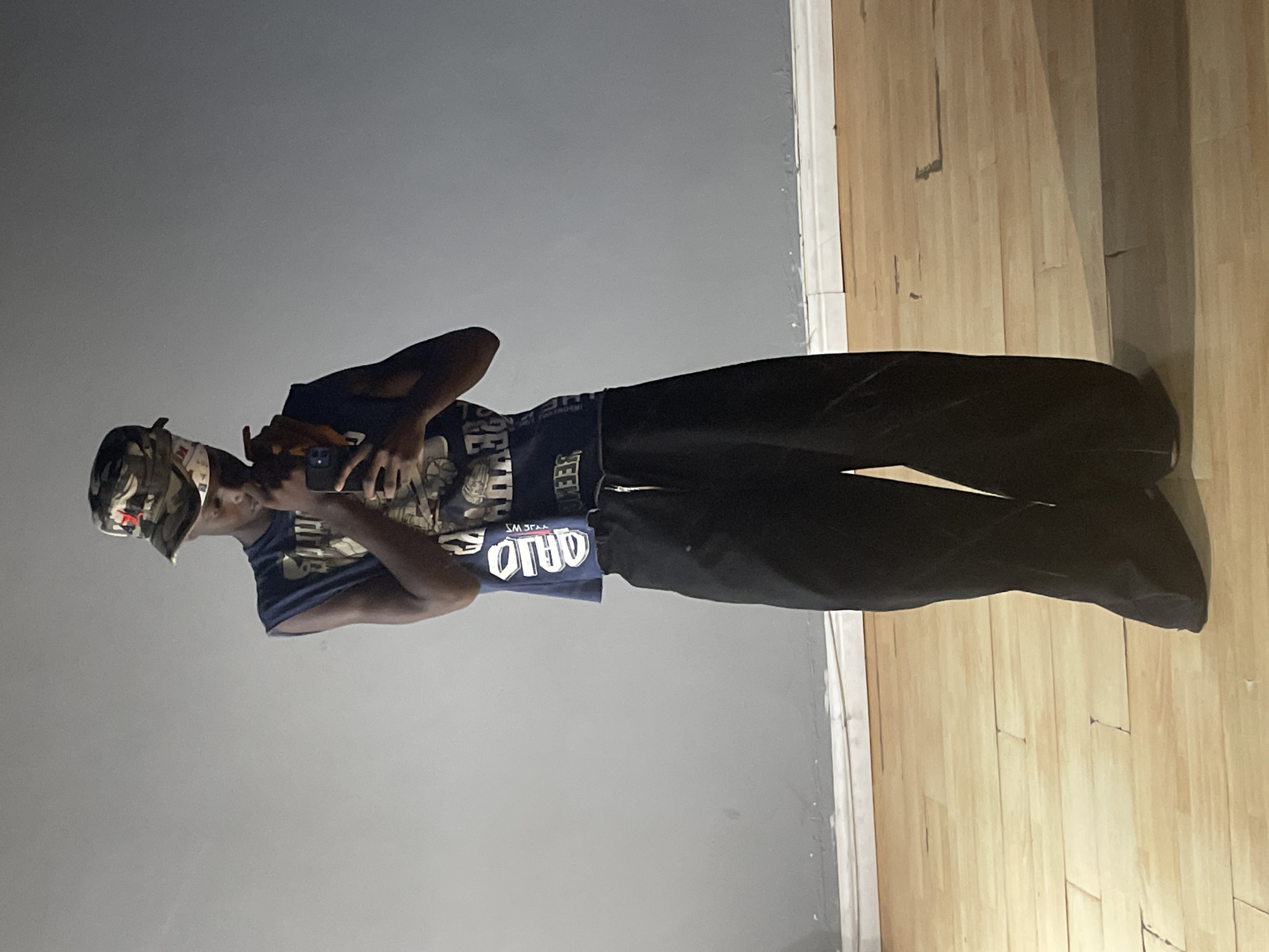 An indoor, waist-up shot captures a person, likely male, standing in a casual pose. They are wearing a camouflage bucket hat, a blue t-shirt with graphics, and dark, wide-leg pants. The person holds a smartphone up with both hands, appearing to be taking a selfie or recording a video, as suggested by the reversed text visible on their t-shirt. The background consists of a plain light gray wall and a light-colored wooden floor.
Visible text on the blue t-shirt includes "OHIO" which appears backward, confirming the selfie perspective, and partial letters like "JEE" and "WS" (or "JEWS") which could be part of a larger design or brand name. The person's face is mostly obscured by the phone and hat, making their expression undiscernible. The setting is simple, implying an everyday moment. The location context indicates this image was taken in Lekki, Nigeria.