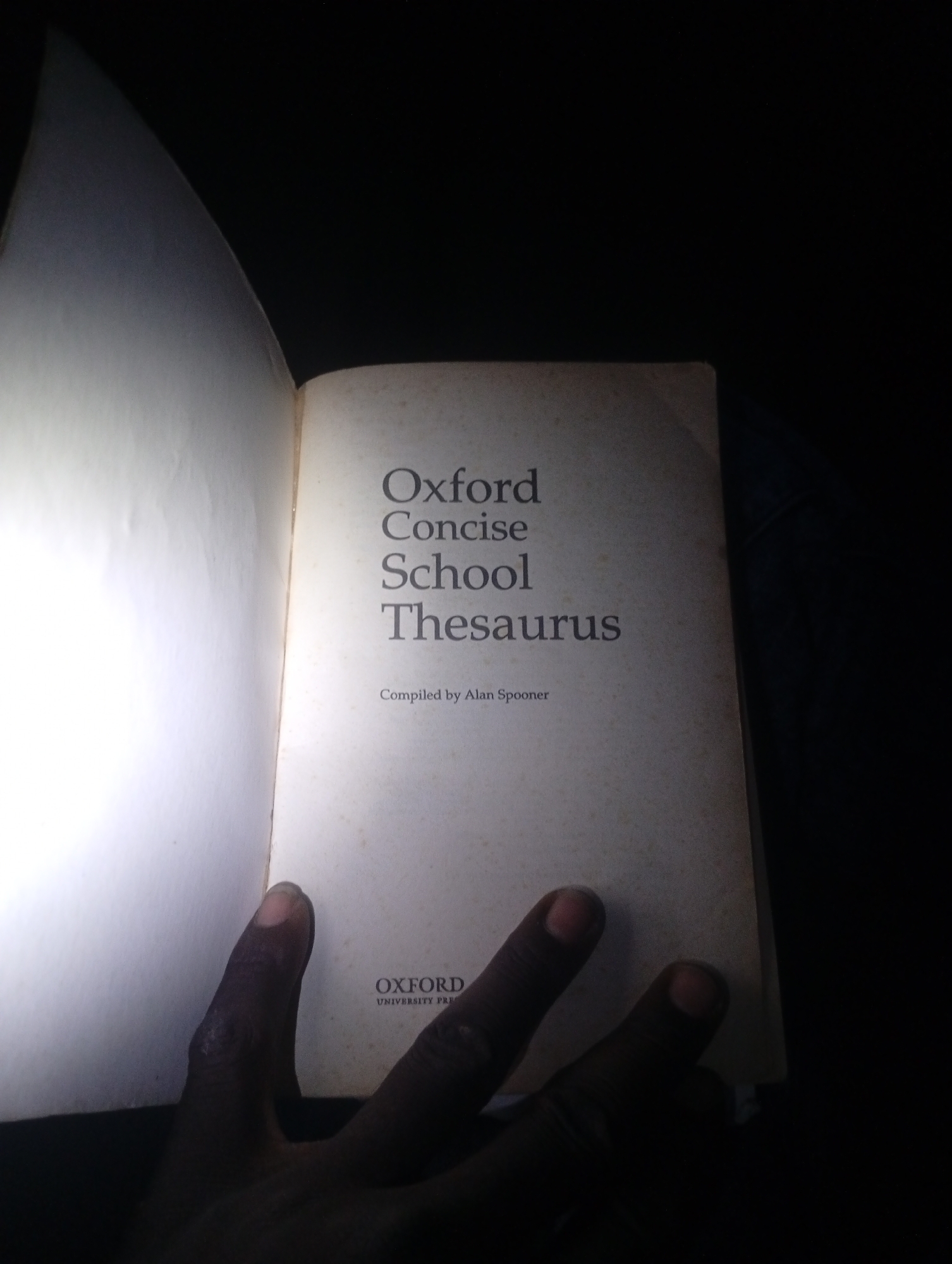 An open book titled "Oxford Concise School Thesaurus" is held by a person's hands in the city of Gulak, Nigeria. The book is positioned on a dark surface, possibly a table or floor, with significant shadows obscuring the environment. The left page of the book is brightly illuminated, contrasting with the right page which displays the title and a smaller inscription reading "Compiled by Alan Spooner." Below this, the "OXFORD UNIVERSITY PRESS" logo is visible, albeit partially obscured by a thumb. The hands appear to be dark-skinned, with visible fingernails. The image is taken from a slightly elevated, forward-facing perspective.