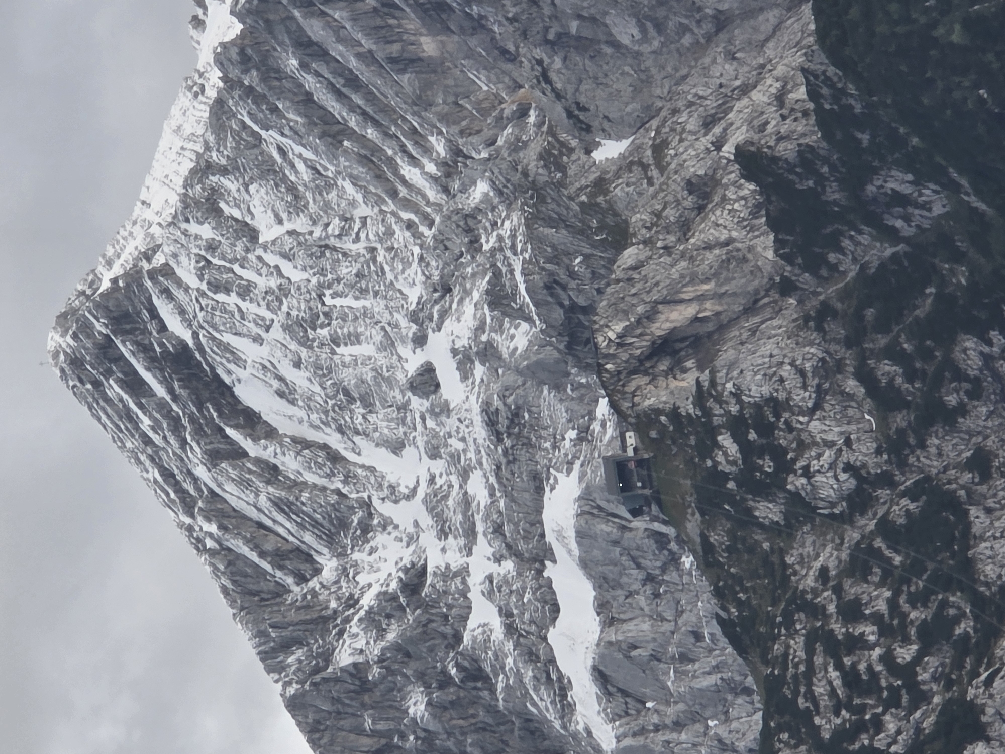 The image captures a close-up view of the Zugspitze, Germany's highest peak, located near Garmisch-Partenkirchen. The mountain's summit is partially covered in snow, with rugged, gray rock formations dominating the landscape. A modern building, possibly a cable car station or a mountain hut, is situated on a rocky outcrop partway up the mountain. The sky above is overcast, casting a muted light over the scene. The lower slopes of the mountain are covered in dark green vegetation.