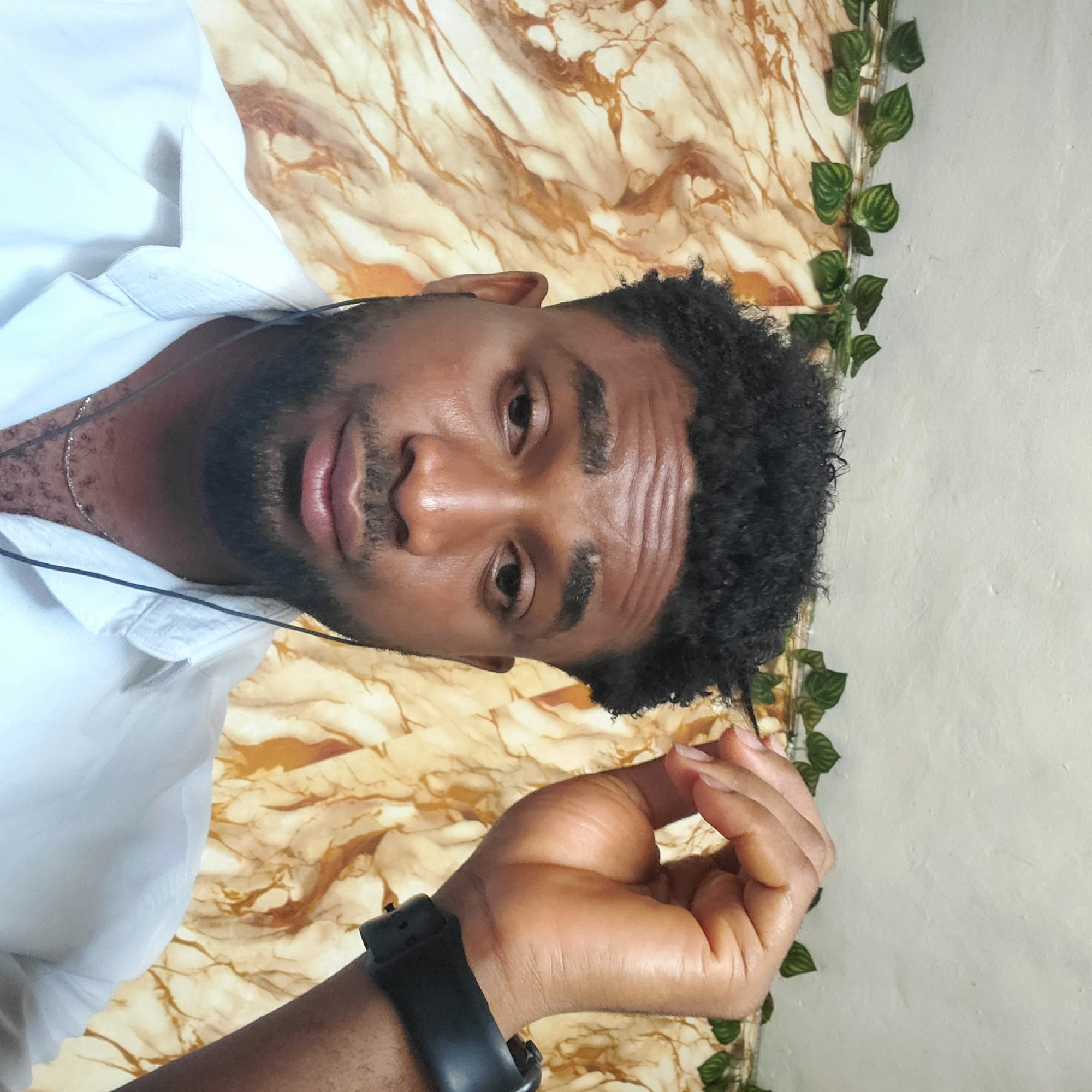 A mid-shot captures a man with dark skin, a beard, and short, curly black hair. He is wearing a white button-down shirt and appears to be in his 20s or 30s. The background consists of a marble-like patterned wallpaper and a decorative green vine with leaves. The man is wearing a black wristwatch and has what appears to be a silver necklace. His face is clean-shaven except for the beard, and he looks directly at the camera. The environment suggests a home or indoor setting. There is no explicit evidence to suggest the location in the city of Nkpor, Nigeria.