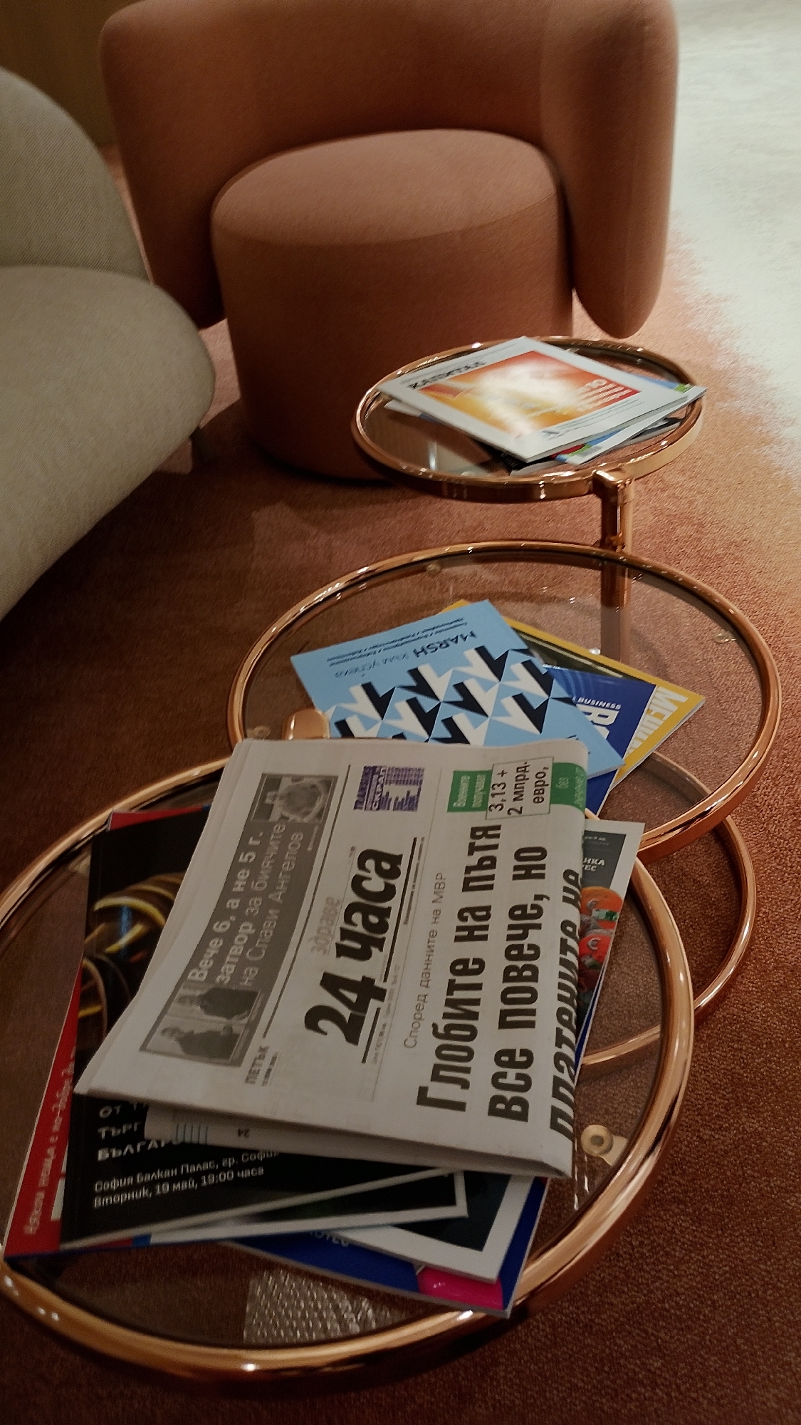An indoor scene depicts a circular, glass-topped side table with a rose gold frame, holding several newspapers and magazines. The table is positioned on a brown carpet. In the background, a section of a cream-colored armchair and a terracotta-colored ottoman are visible. One newspaper, with the masthead "24 ЧАСА," is prominently displayed. Other visible text includes "България" and "София балкан Палас, гр. София." The overall impression is of a reading area within a modern, upscale establishment.