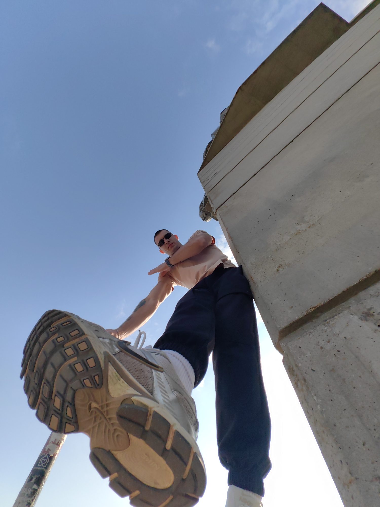 A low-angle shot depicts a man from below, with a foot in the foreground, as if about to step on the viewer. The man is wearing sunglasses, a light-colored t-shirt, and dark trousers. He has a tattoo on his arm and is pointing his index finger. His shoe is a light-colored athletic shoe with a worn sole. Behind him is a large concrete structure, possibly a bridge support, set against a clear blue sky. The photograph appears to be taken outdoors in a sunny location, and while the exact location in Shumen, Bulgaria is difficult to determine from the image alone, the concrete structure suggests an urban or industrial setting within the city.