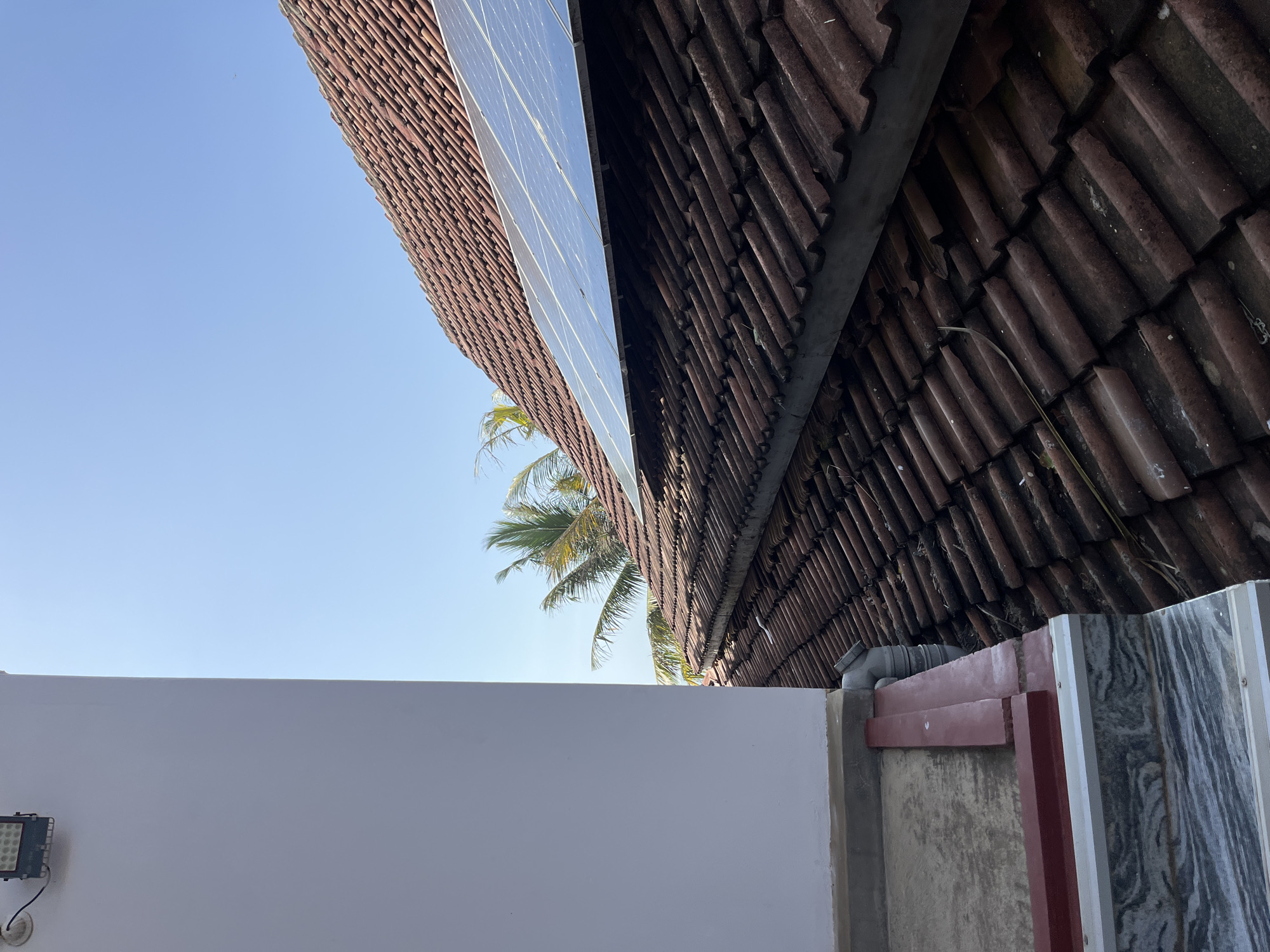 The image shows a close-up view of a tiled roof with solar panels installed on a sunny day. The tiles are reddish-brown and appear weathered. The solar panels are rectangular and reflect the blue sky. In the foreground, a portion of a white wall and a concrete ledge are visible. The scene is set in Matara, Sri Lanka, as indicated by the presence of palm trees in the background, typical of the region's tropical climate. The sky is clear and blue, suggesting good weather conditions. No people are visible in the image.