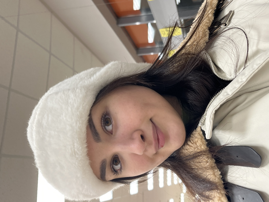 A female individual is depicted in a close-up, eye-level photograph, positioned indoors in the city of Neftekamsk, Russia. She has long, dark brown hair, brown eyes, and fair skin, with a small dark mole or blemish visible on her upper lip, to the right of the philtrum. She is wearing a white faux fur hat, a dark green turtleneck, and an open, light-colored (beige/cream) jacket featuring a prominent tan faux shearling collar. A metallic zipper and a dark brown strap are visible on the jacket's front. The subject displays a subtle, closed-mouth smile and looks directly at the camera. The background consists of a brightly lit interior space with white square-panel ceiling tiles and overhead fluorescent lighting fixtures. To the right, an orange wall or panel is visible above a grey structural element, possibly a window frame or partition, beyond which a reflective surface is present.