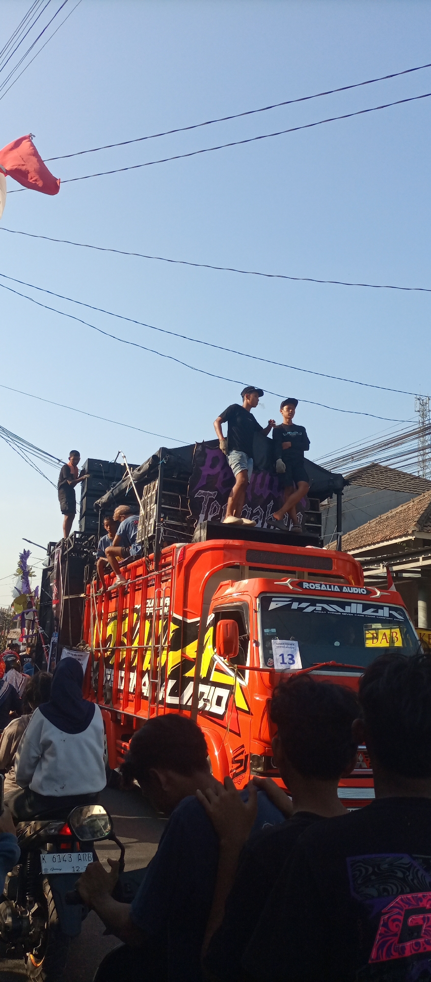 This image depicts a street scene in Jekulo, Indonesia, likely during a daytime event or parade. A prominent feature is a large, brightly painted orange truck with a sound system and speakers mounted on its flatbed. Several young men are visible on top of and around the truck. Two men in black t-shirts and hats are standing on the speaker setup, with one appearing to smoke. Another man in a black t-shirt is standing to the left of the truck. In front of the truck, the side is adorned with large yellow and black graphics.
A crowd of people is gathered on the street, with some individuals on motorcycles. In the foreground, several people are seen from behind, including a person wearing a blue hijab and a white jacket, and others in dark clothing. One person is seated on a motorcycle, facing away from the camera, and their motorcycle's license plate is visible, reading "K 6143 ARB". The sky above is clear and blue, with several power lines crisscrossing the scene. The background includes buildings and trees, suggesting a typical urban or semi-urban environment.