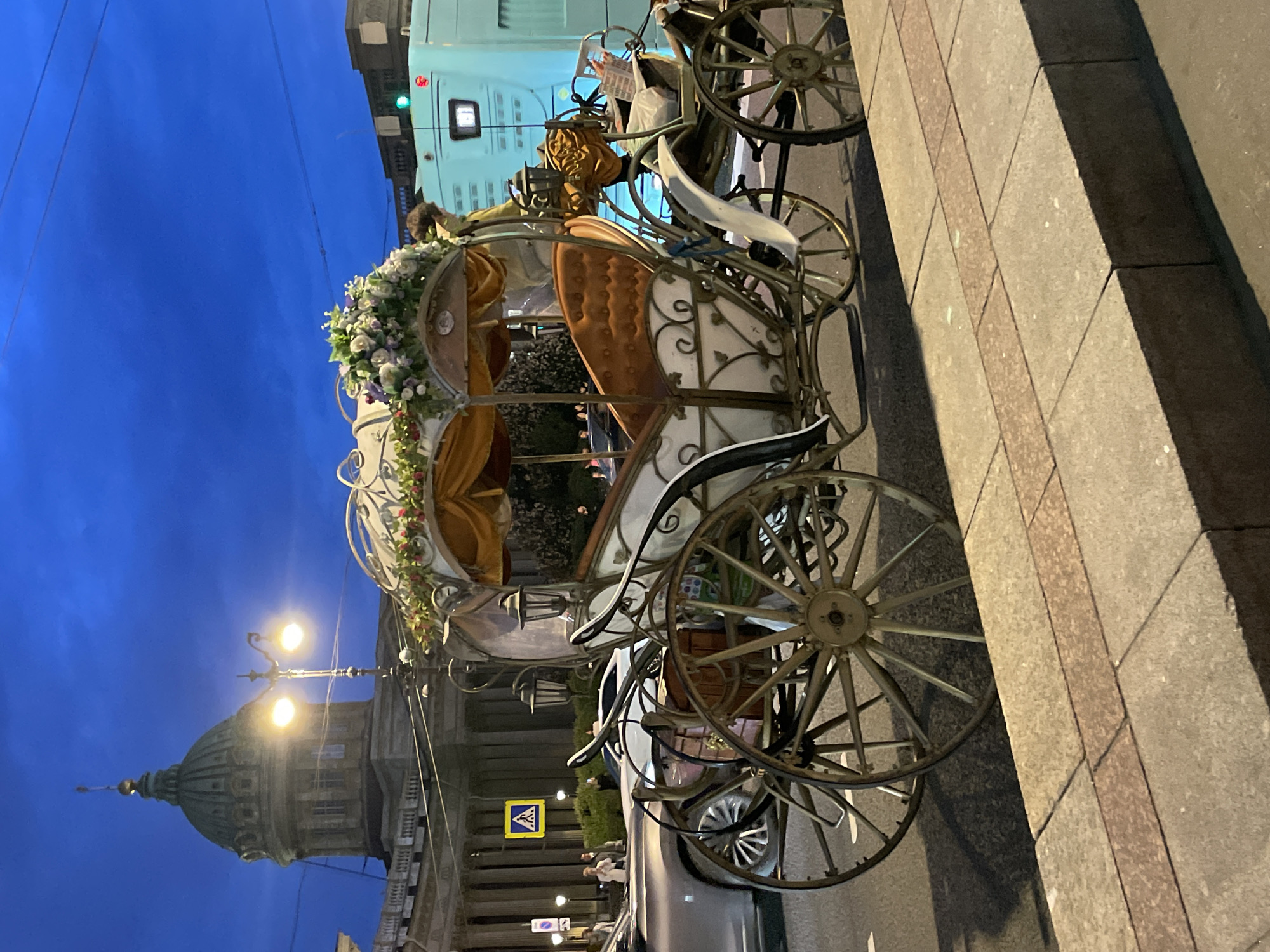 The image captures an ornate, white and gold carriage decorated with flowers, parked on a street at dusk. The carriage features plush orange seating and intricate metalwork. Behind the carriage, a turquoise bus is visible, and in the background, a large building with a dome, resembling a cathedral, stands under a dark blue sky. Streetlights illuminate the scene, casting a warm glow on the cobblestone sidewalk in the foreground. The environment suggests an urban setting, likely a tourist area, given the presence of the carriage.