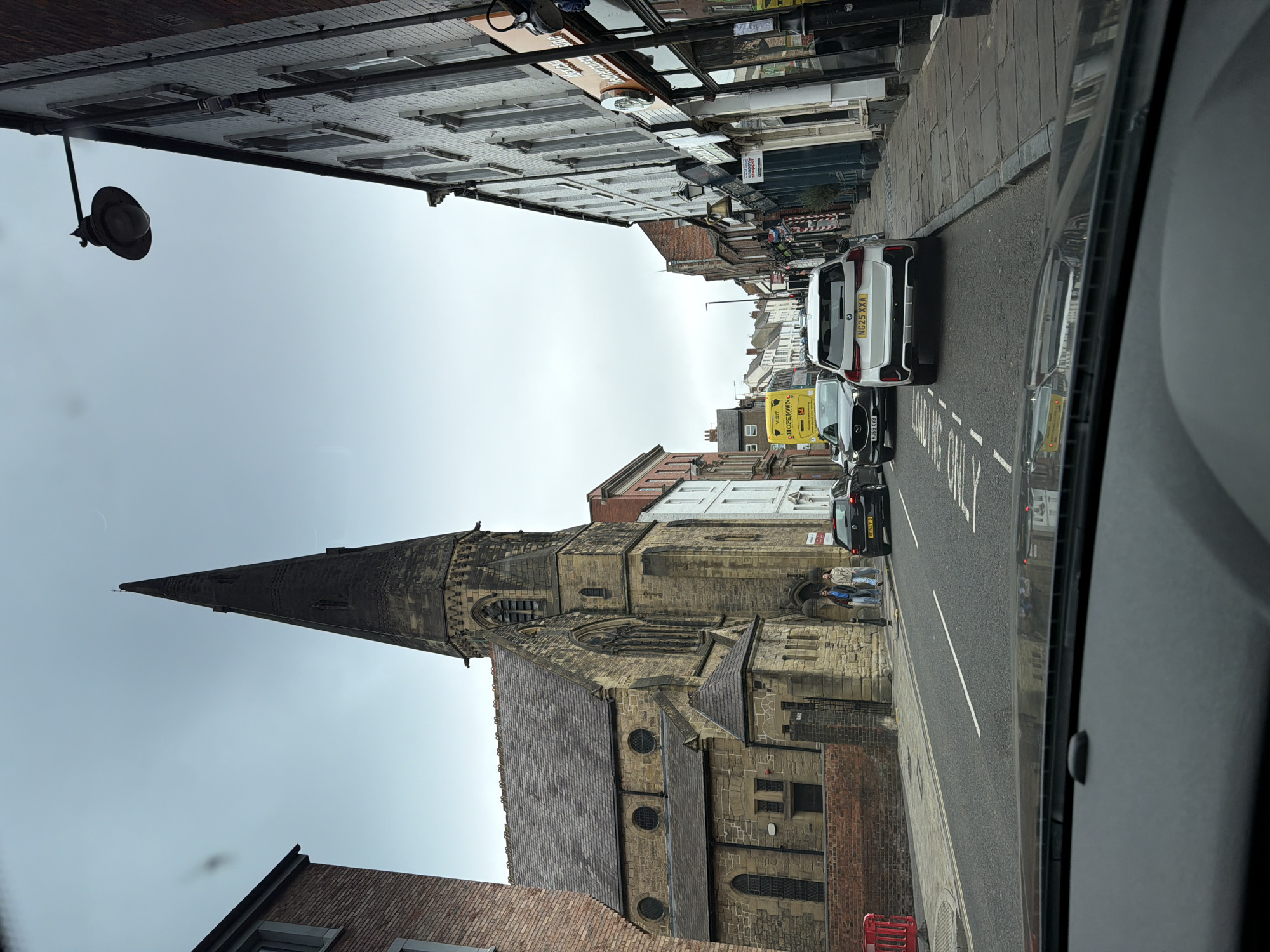 Captured from inside a vehicle, the photograph depicts a street scene in Durham, United Kingdom, angled slightly upwards to capture the cityscape. Prominently featured on the left is the Gothic architectural style tower and structure of St. Mary-le-Bow church. A wet or recently wet road surface indicates recent precipitation. On the right-hand side of the road are parked and moving vehicles, including a white BMW with license plate NG25 XXA, and a black vehicle with license plate ending in RXRE. Pedestrians are visible alongside the church. The sky is overcast. The buildings lining the street vary in architectural styles and heights, and feature storefronts on street level. The image appears to have been taken while the vehicle was stationary or moving at low speed.