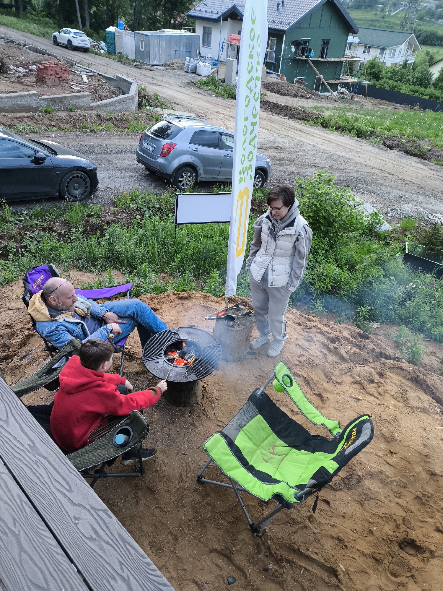 The image shows an outdoor scene featuring a family gathered around a fire pit. A man sits in a purple camping chair, wearing a blue denim jacket and jeans. A boy in a red hoodie sits in a green camping chair next to the fire pit, holding a stick and seemingly tending to the fire. A woman stands nearby, wearing a light-colored vest and pants, with her hands in her pockets. Smoke rises from the fire pit, which sits on a dirt or sandy surface. An empty green camping chair lies on the ground nearby. A white banner with green writing is behind the woman.
In the background, two cars are parked in a dirt lot, and a partially constructed house is visible with workers on scaffolding. The location appears to be a construction site or a residential area under development. A portable toilet and some construction materials are also visible in the background.