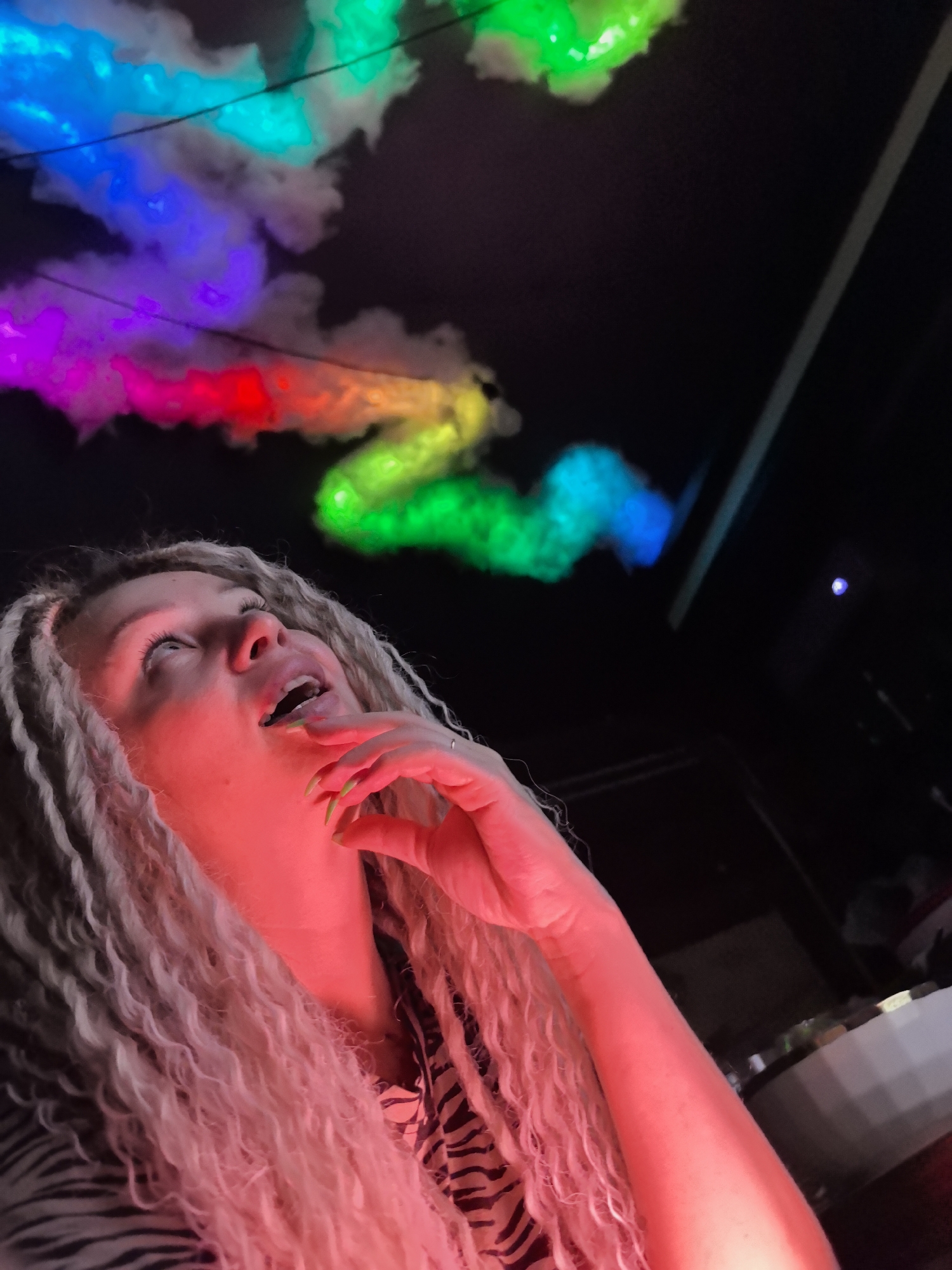 A woman with long, light-colored, curly hair looks up at a ceiling adorned with colorful, cloud-like decorations lit with vibrant hues of red, orange, yellow, green, blue, and purple. Her right hand is near her face, and her mouth is open. The woman is wearing a patterned shirt. The background is dark, with a few indistinct objects visible. The woman and the clouds appear to be illuminated by a red light. The city of Noviy, Russia is not discernible from the image.