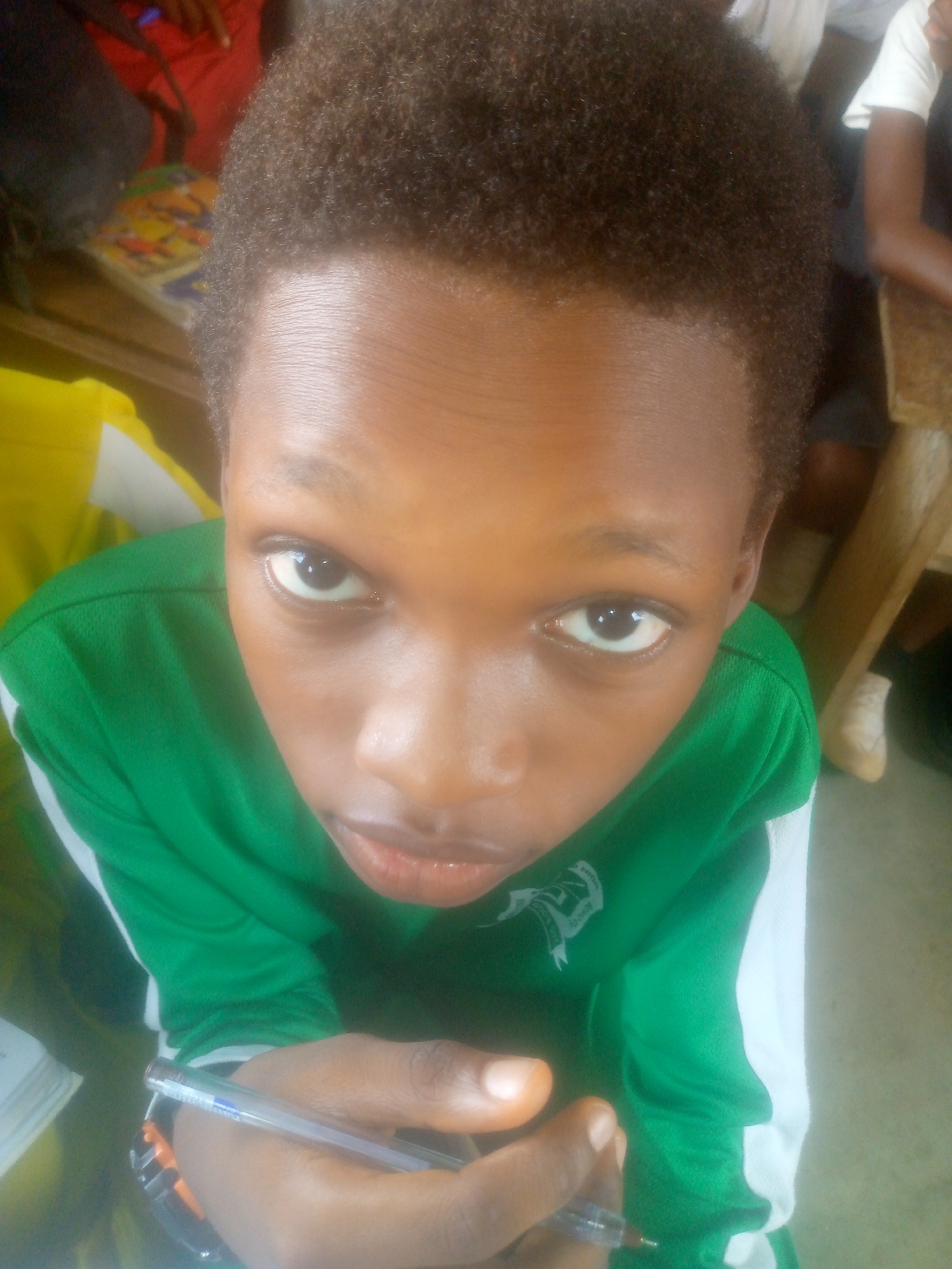 A close-up, upward-angled shot captures a young person in Unknown, Unknown. The individual, likely a student, is wearing a green shirt with a white stripe along the shoulder, and a visible emblem on the chest. They hold a pen in their hand. The setting appears to be a classroom or similar environment. Other people and objects are partially visible in the background, including chairs and a book. The location is generally indistinct.