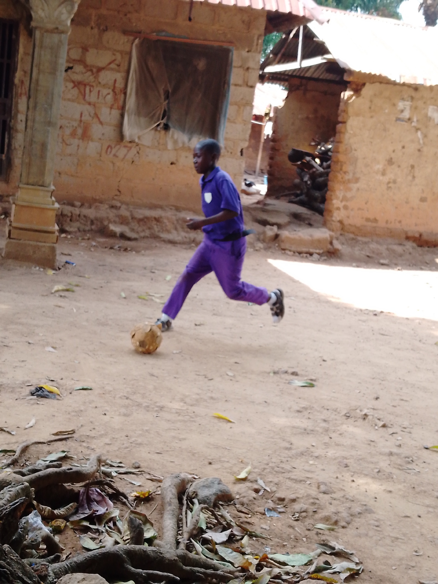 A young male is dribbling a ball in a village. The boy is wearing a purple shirt and pants with black and white shoes. He appears to be running. The area where he is playing has a dirt surface with exposed roots in the foreground. In the background are buildings constructed with mud bricks and metal roofs. A collection of firewood is seen piled near the building on the right. The location is Unknown, Unknown.