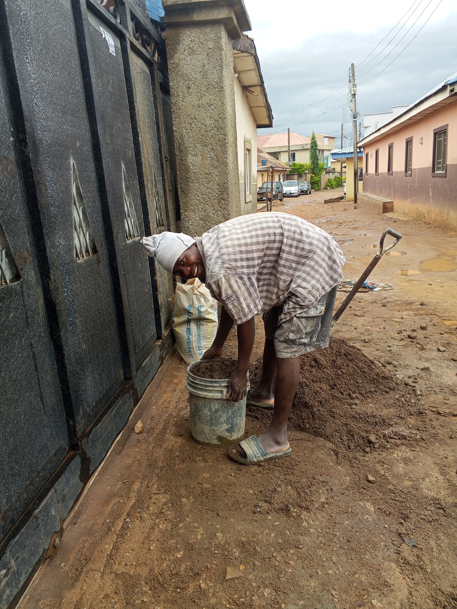 The image captures a person engaged in manual labor on what appears to be a residential dirt road on an overcast day.
The primary subject is a person, likely male, dressed in a plaid short-sleeved shirt, dark cargo shorts, a light-colored head covering, and striped slide sandals. They are bent over, actively scooping dark, moist earth or sand from a pile into a gray plastic bucket. The person is smiling broadly and looking towards the camera. A shovel is leaning against the pile of earth, and a white sack, which appears to be a cement bag with the word "CEMENT" clearly visible, rests behind them near a large, dark metal gate.
The scene is set outdoors in what looks like a less developed urban or a rural residential area. To the left, a tall, dark metal gate with a textured, possibly hammered, surface runs along the property line, adjacent to a beige textured pillar. The ground is unpaved, consisting of reddish-brown dirt, with several muddy patches and small puddles, suggesting recent rain or damp conditions.
In the background, a street extends with various one-story houses flanking it. Some houses are painted light beige, while others feature a distinctive reddish-brown lower half and a lighter upper section. Several cars are parked along the dirt road. Utility lines crisscross the sky, and a utility pole equipped with a loudspeaker stands prominently in the middle distance. The sky is uniformly overcast.
Notable details include the person's positive demeanor despite the labor, the wet and muddy ground, and the basic infrastructure of the street. The visible text on the white sack clearly reads "CEMENT," with additional, less legible text that might be "BUOCEMENT" above it.