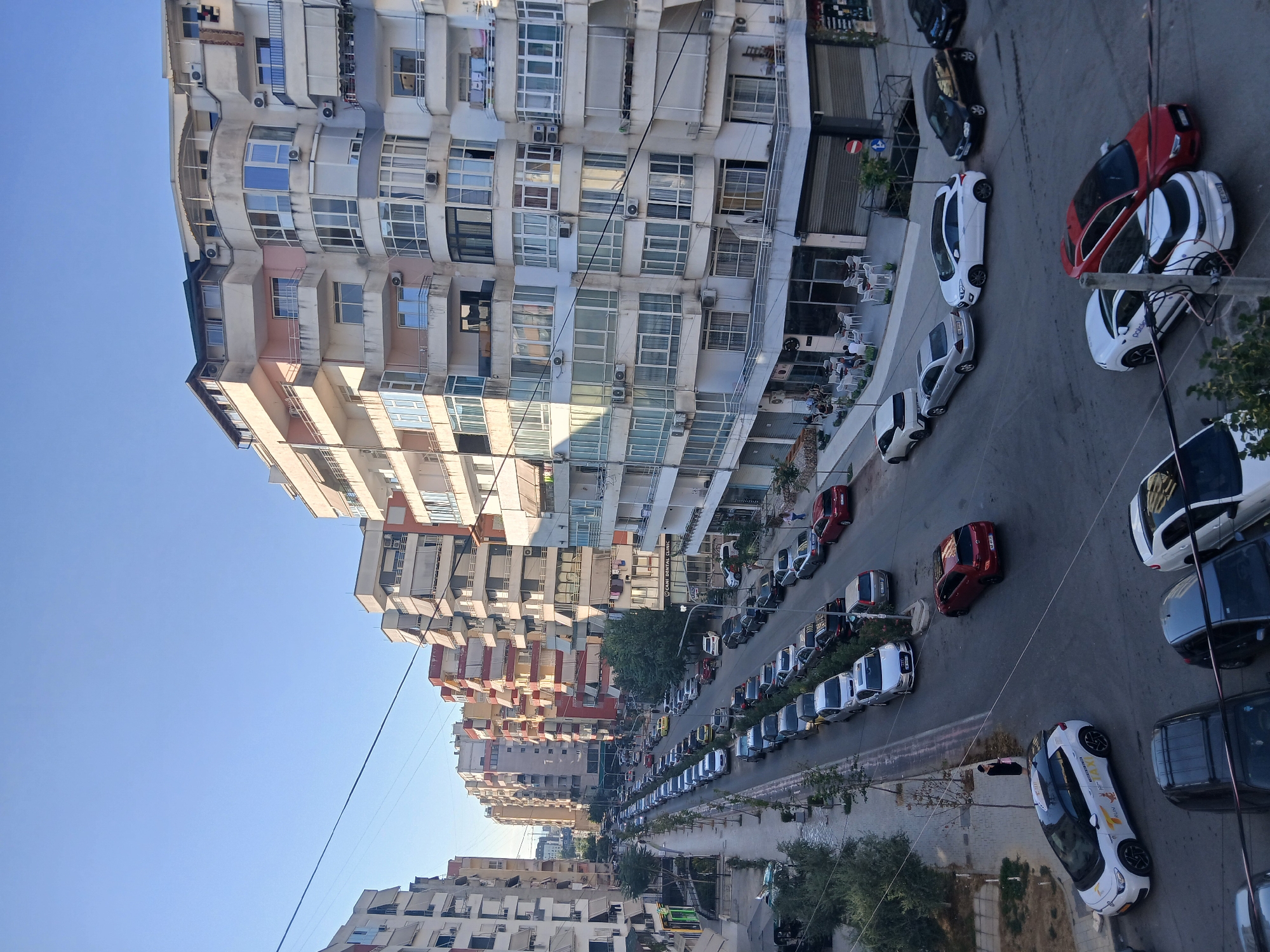 This image depicts a street scene in Mëzez, Albania, characterized by multi-story apartment buildings lining both sides of a road filled with parked and moving vehicles. The buildings exhibit a range of architectural styles, with many featuring balconies, large windows, and a mix of light-colored facades, some with accent colors like pink and yellow. The street itself is asphalted and appears to be a main thoroughfare, with lanes designated for traffic and parallel parking.
A significant number of cars are parked along the street, predominantly in a single file on the right side and a double file on the left. The vehicles are of various makes and models, including sedans, SUVs, and what appears to be a white electric taxi with prominent branding. A few cars are in motion, suggesting active use of the street.
On the sidewalk to the left, there are trees and some low-lying vegetation. Several shops and cafes are visible at street level, with outdoor seating areas occupied by a few individuals. A person in dark clothing stands on the left sidewalk near a parked vehicle. The sky is clear and blue, indicating daytime, and the sunlight casts shadows, suggesting it is either morning or late afternoon. The overall impression is a busy urban environment, typical of a residential and commercial district.