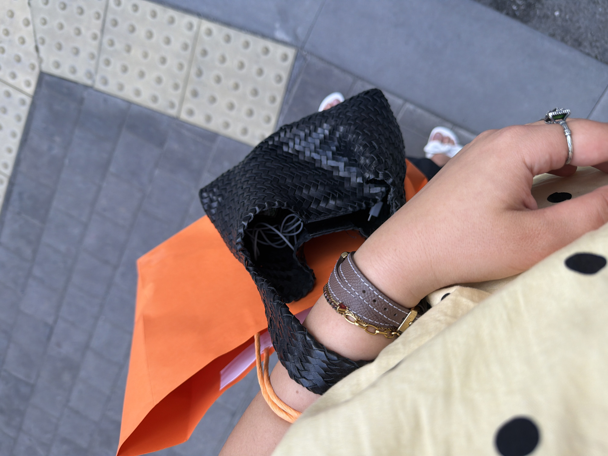 The image captures a top-down perspective of a person walking on a sidewalk in Yerevan, Armenia. The sidewalk is composed of grey rectangular pavers with a section of textured beige pavers with circular indentations to the left. The person is wearing a light yellow dress with large black polka dots.
On the person's arm, there are several bracelets: one light brown leather bracelet, a gold chain bracelet, and an orange cord bracelet. A silver ring with a green stone adorns the person's finger.
The person carries a black woven bag and an orange paper bag. Partially visible are the person's feet in white sandals.