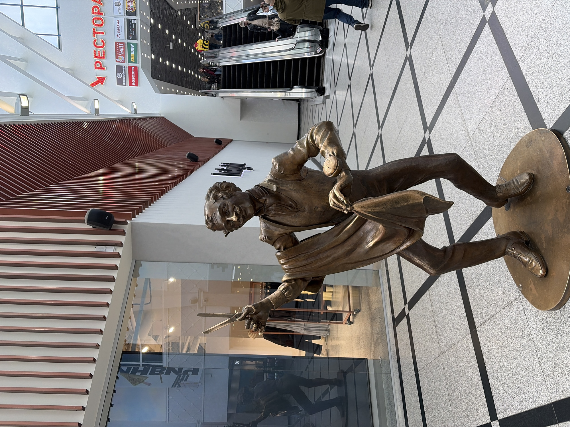 The image shows a bronze statue of a man in period attire, holding a fencing foil and mask, set on a circular bronze base within a public space. The location appears to be inside a modern shopping mall or similar complex, with an escalator visible in the background and a "РЕСТОРАН" (RESTAURANT) sign in red Cyrillic letters. The floor has a gray and black tiled pattern. Reflections in nearby glass suggest retail store interiors. The statue is located in Yekaterinburg, Russia.