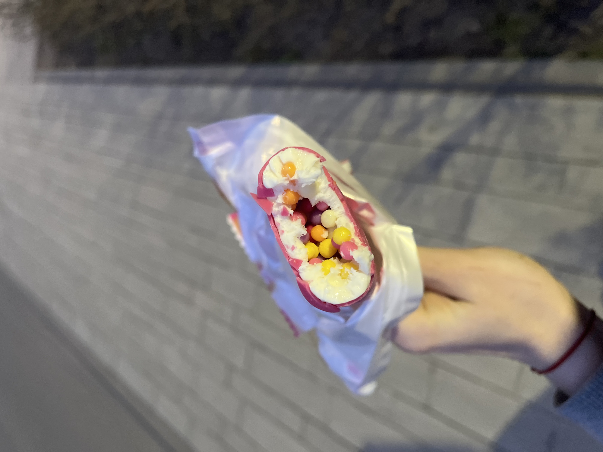 A hand holds a partially eaten, brightly colored dessert in a white wrapper outdoors, likely on a street or sidewalk. The dessert appears to be cylindrical, with a pink exterior and a white filling speckled with multi-colored candies or balls. The hand is fair-skinned and partially visible, with a red string or bracelet around the wrist. The background shows a blurred gray surface, possibly a paved area, and some darker out-of-focus elements suggesting foliage or buildings. The image lacks clear markers for pinpointing the specific location in Saint Petersburg.