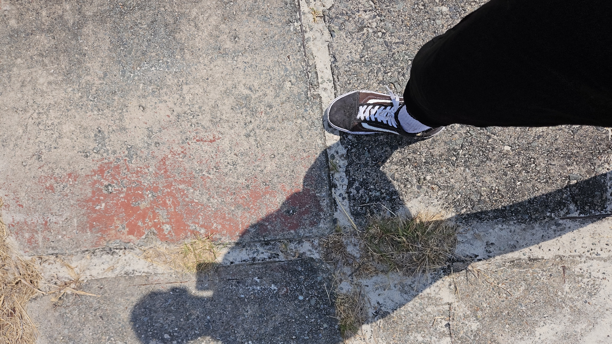 A single foot wearing a black and brown Vans sneaker with white laces and a white sock stands on concrete. The person is also wearing black pants. The concrete appears to be a sidewalk or paved area, with variations in texture and color, and some patches of red. There is some dry grass or weeds growing in a crevice of the concrete. The person's shadow is cast on the concrete. I don't know the location in Cam Ranh, Vietnam.