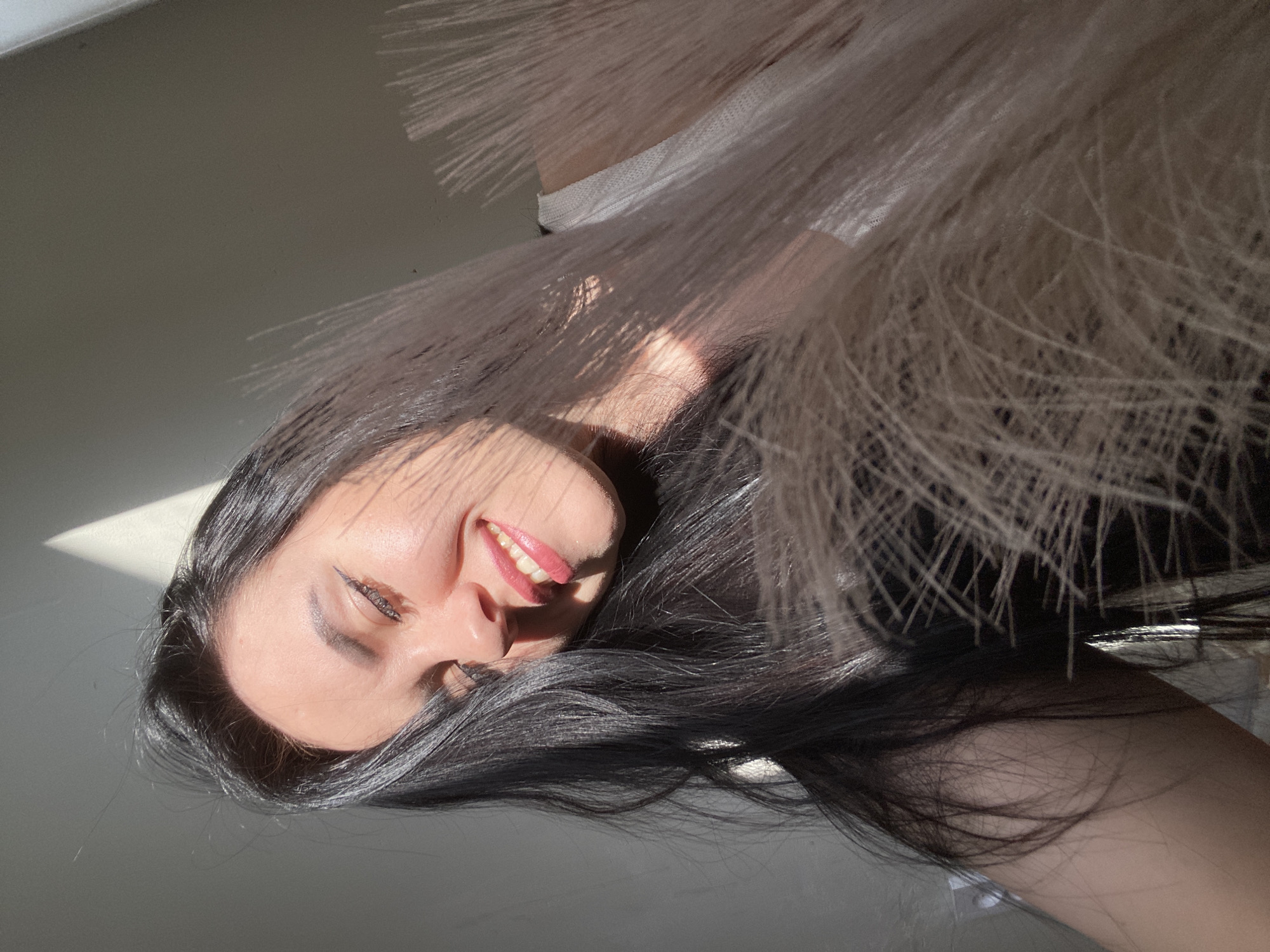 A tilted, close-up portrait captures a smiling young woman with long, dark hair. Her face is illuminated by strong, direct sunlight, creating bright highlights and distinct shadows. She is smiling broadly, revealing her upper teeth, which appear to have braces. Her visible eye shows dark eyeliner.
The woman's dark hair cascades around her face and extends into the lower and left portions of the frame. In the upper right, a mass of feathery, light-colored plumes, possibly pampas grass, adds a soft, decorative element and partially obscures the background.
The background consists of a plain, light-colored surface, likely a wall, onto which the strong sunlight casts a diagonal shadow pattern. The overall lighting suggests a sunny daytime setting. The image evokes a sense of happiness and ease, with the woman's cheerful expression. No visible text is present in the media file. The context for this image is Chaikovsky, Russia.